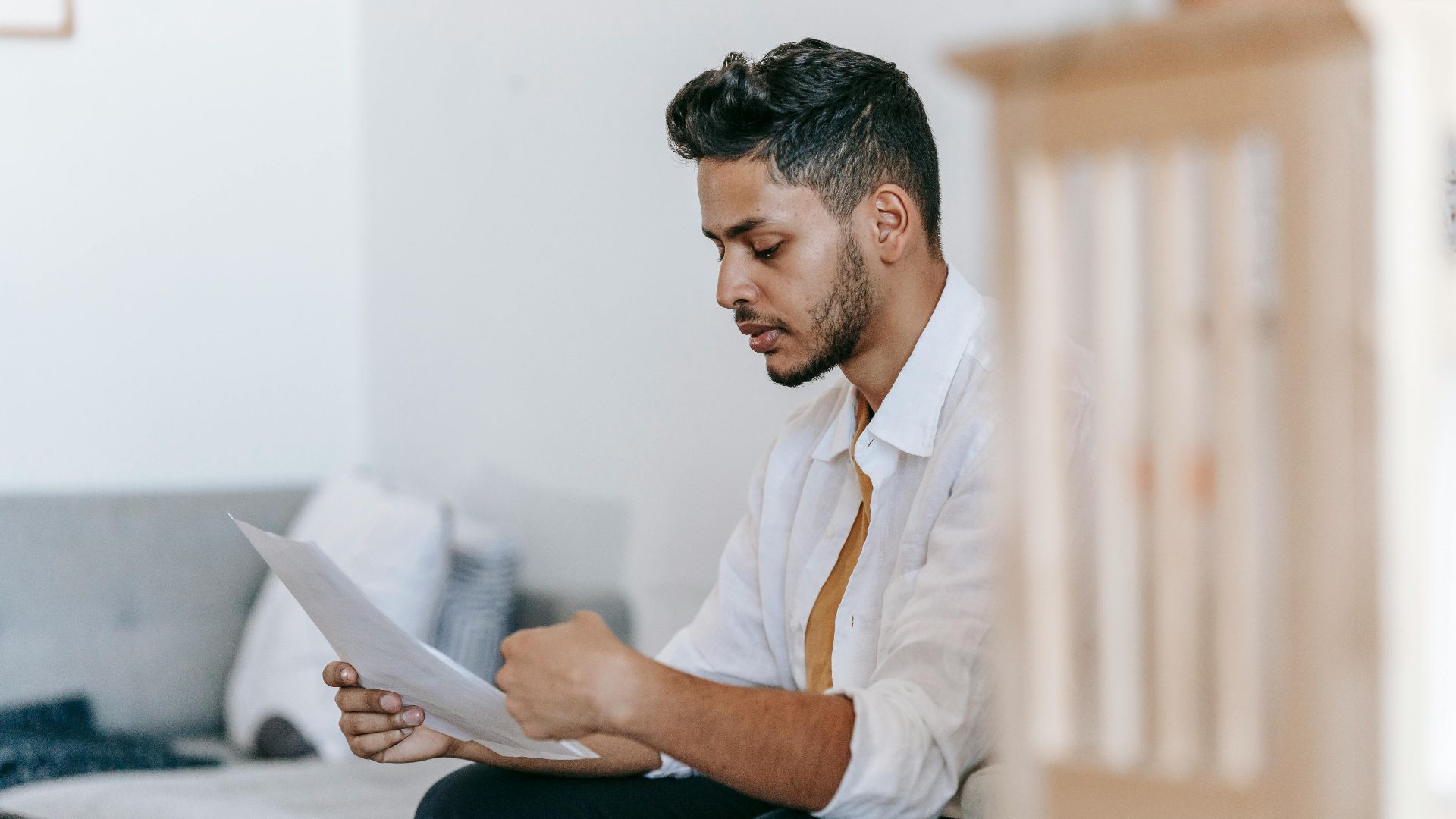Concentrated young ethnic male wearing casual outfit reading documents and sitting on couch in modern living room