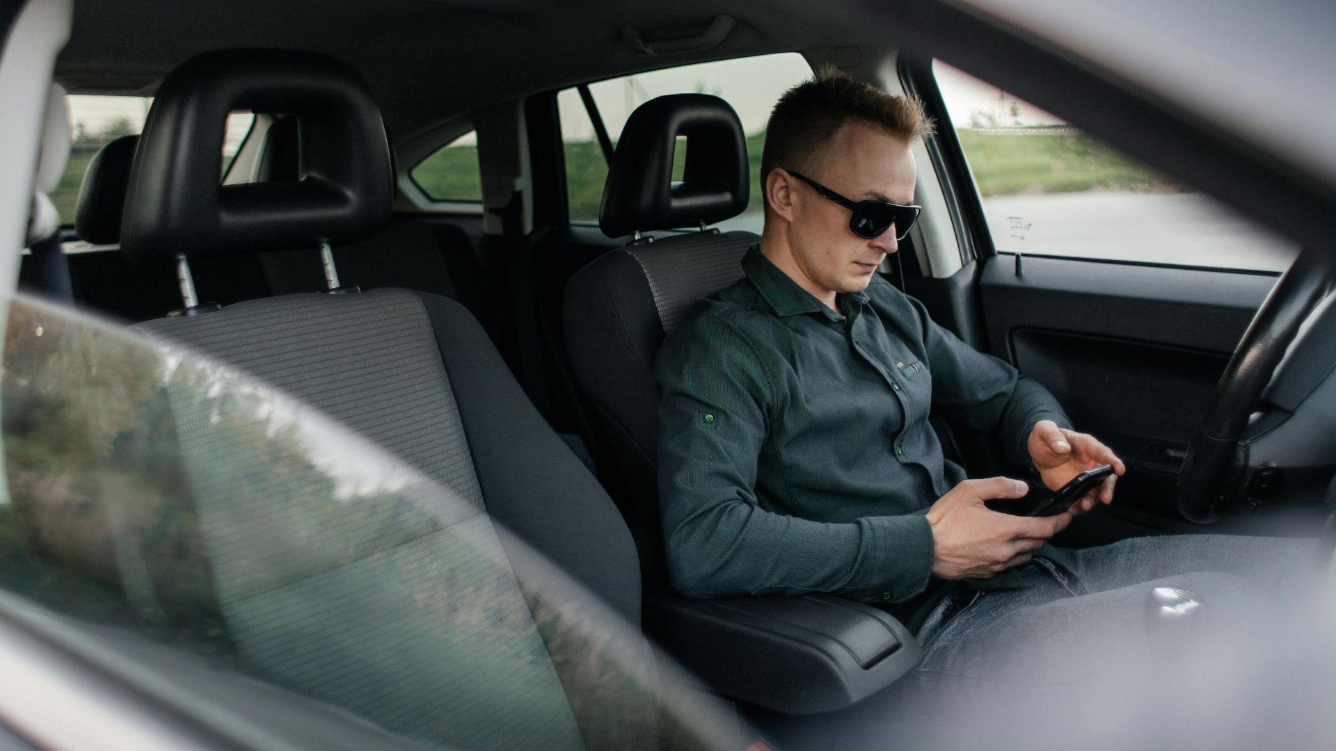 Young man using smartphone inside a parked car, wearing sunglasses.