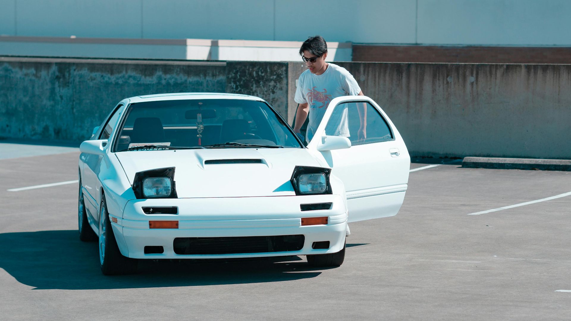 Man getting into a classic white sports car in an empty parking lot under a clear blue sky.