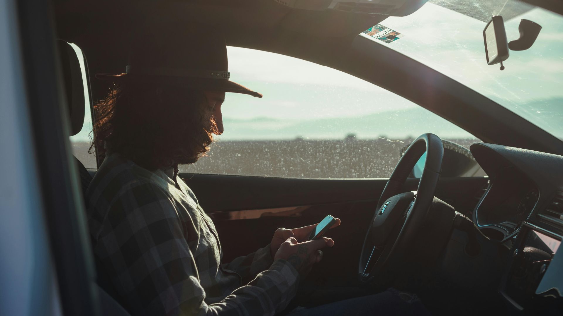 Profile of a man with a hat using a phone inside a car during the day.