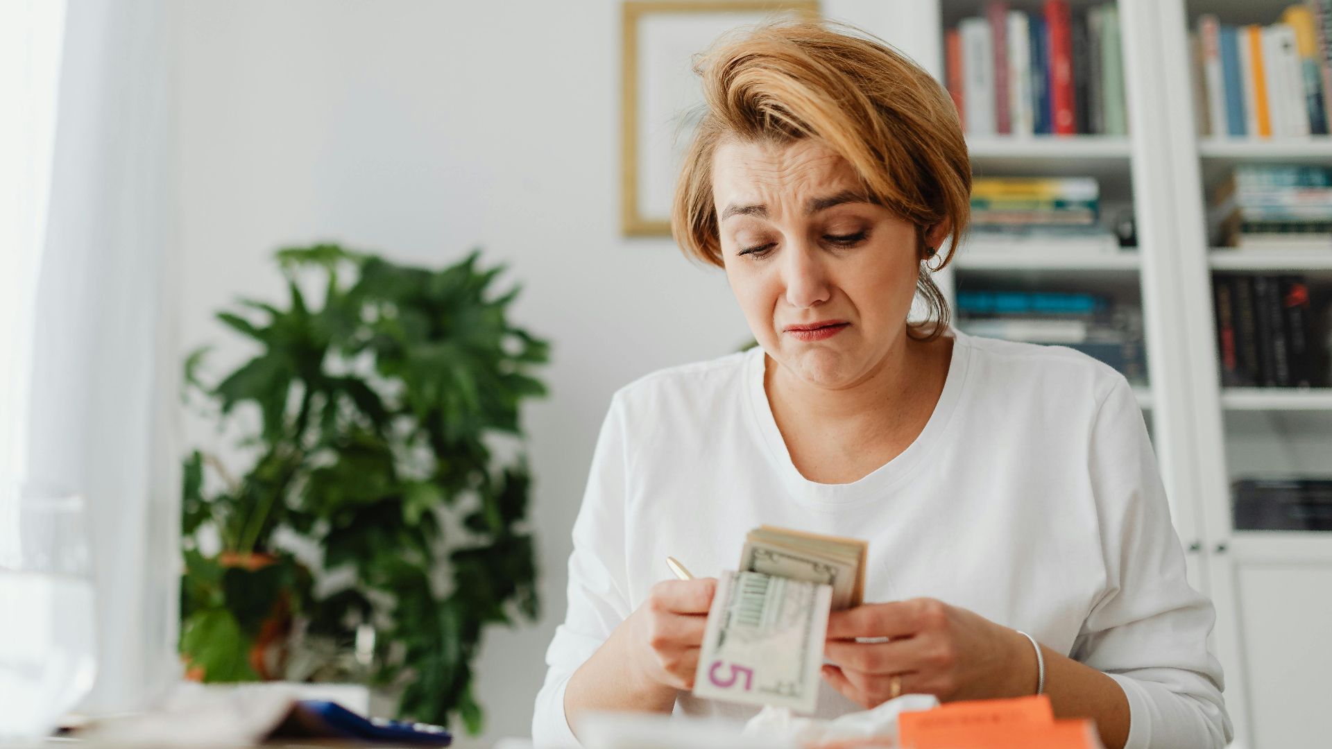A woman concernedly counts money at her desk in a bright room.