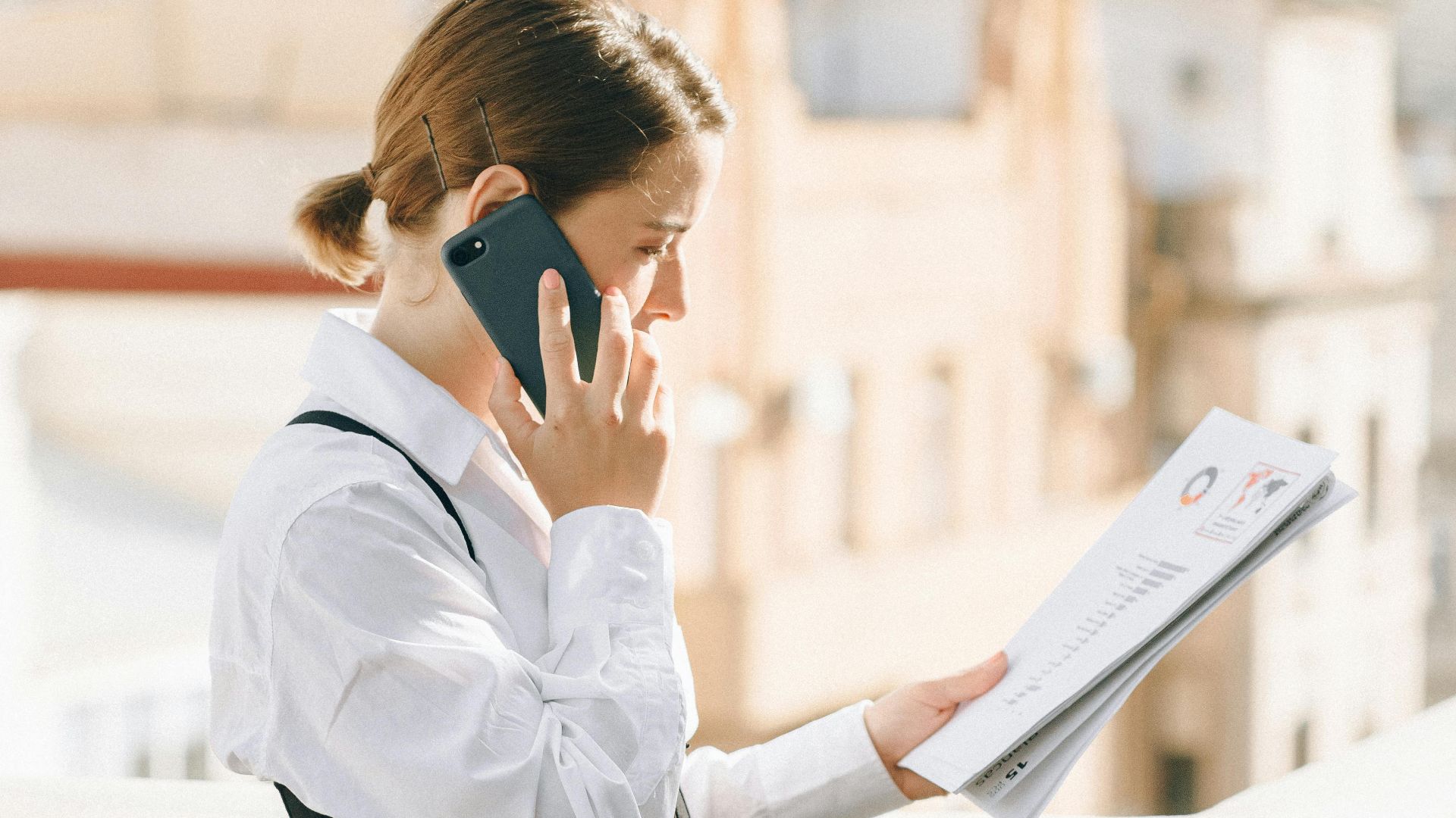 Woman standing on a balcony, holding documents and talking on the phone, wearing a business attire.