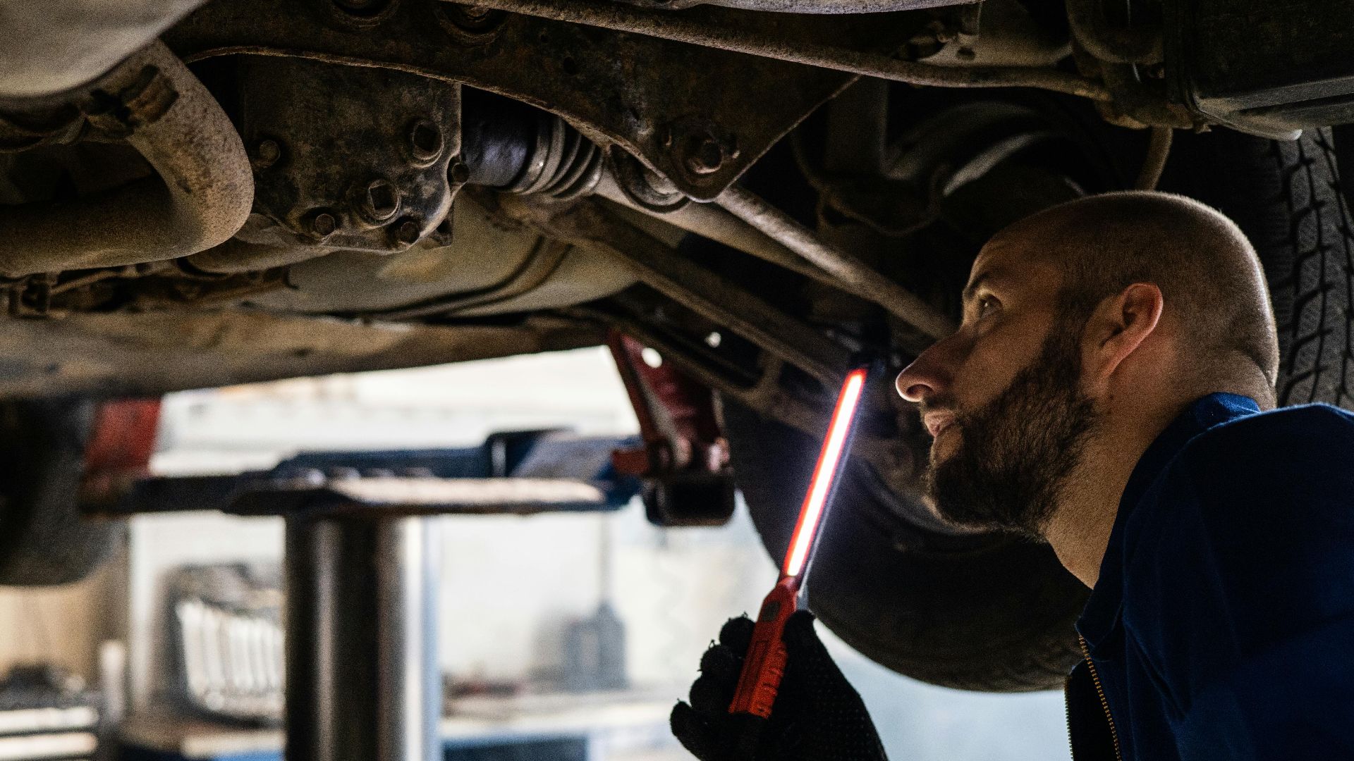 Mechanic using light to inspect vehicle undercarriage in workshop for maintenance.