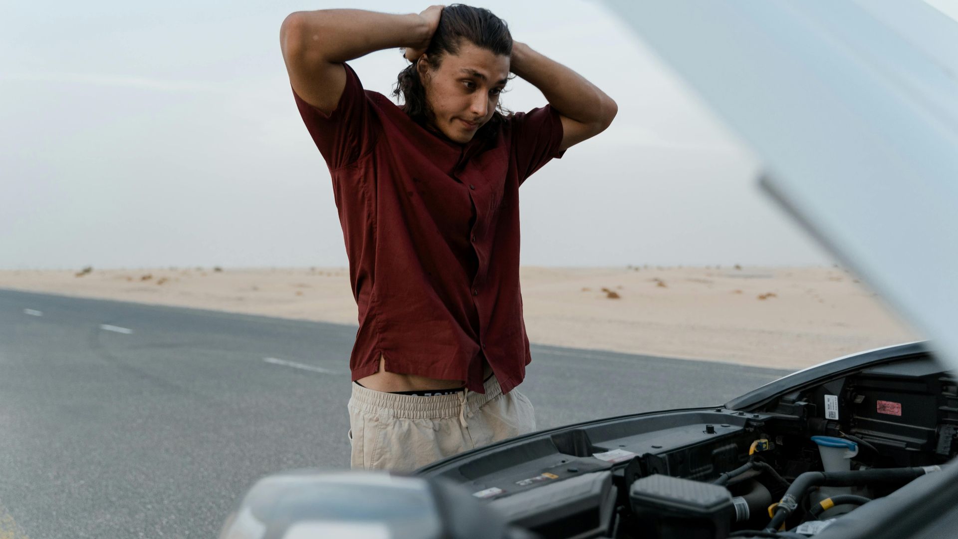 A frustrated man examining a broken car engine on a deserted road, showing stress and concern.
