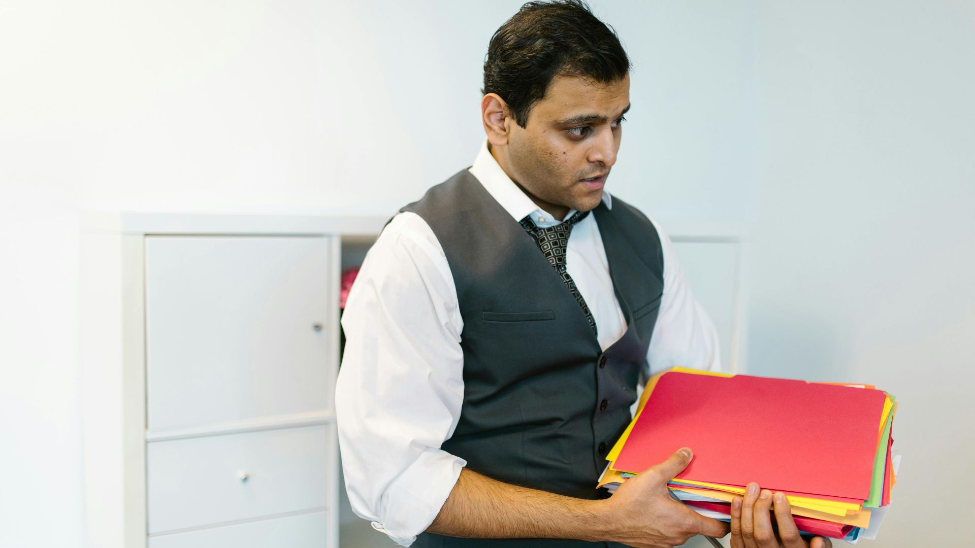 Male office worker in formal attire holding colorful folders in a modern office space.
