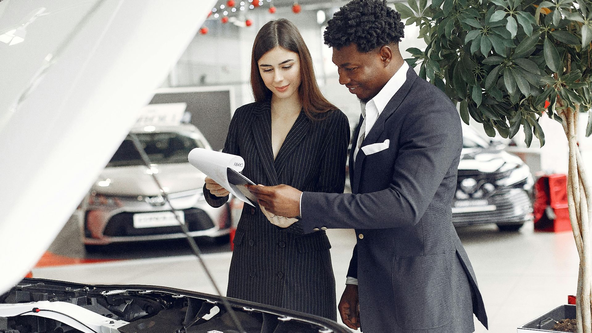 Confident smiling successful black businessman in classy formal suit standing near open motor hood in car showroom and discussing car characteristics with gorgeous female dealer