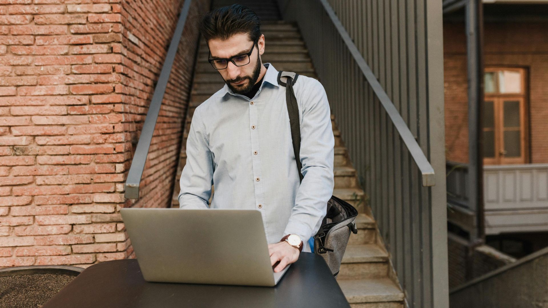 A man with glasses works on a laptop outdoors near brick stairs, depicting remote work lifestyle.