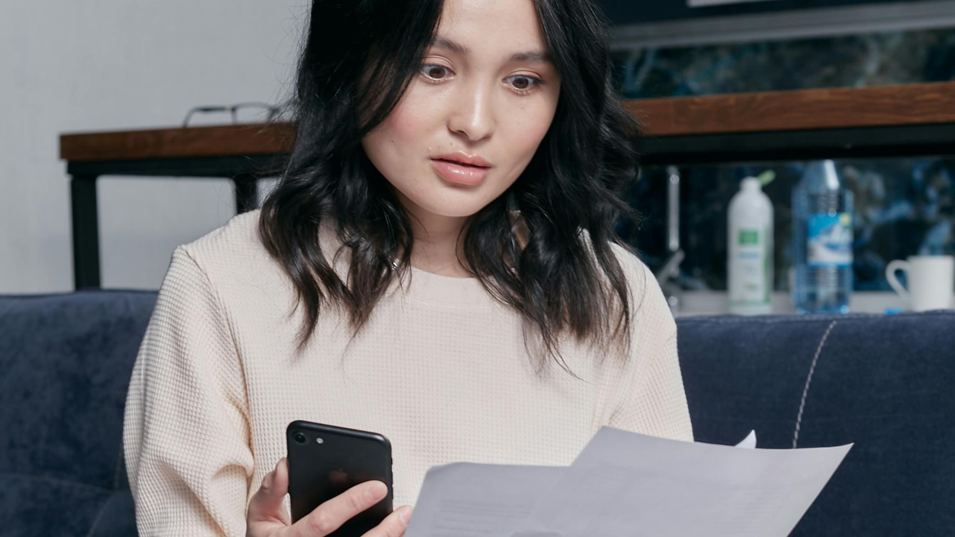 Young woman reviews documents and phone indoors, showcasing concern while seated on a couch with scattered papers. Business concept.
