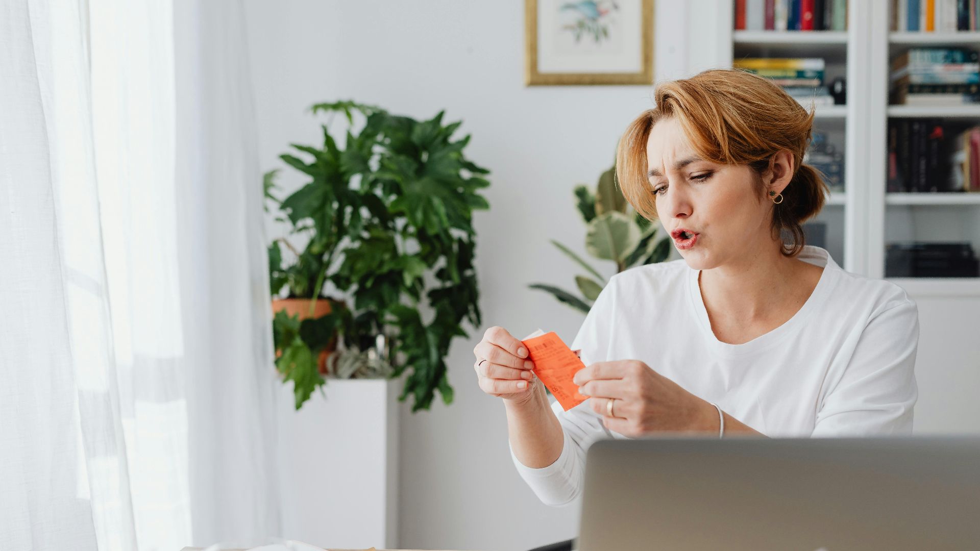 Woman reviewing bills at home desk with laptop and plants, managing personal finances.
