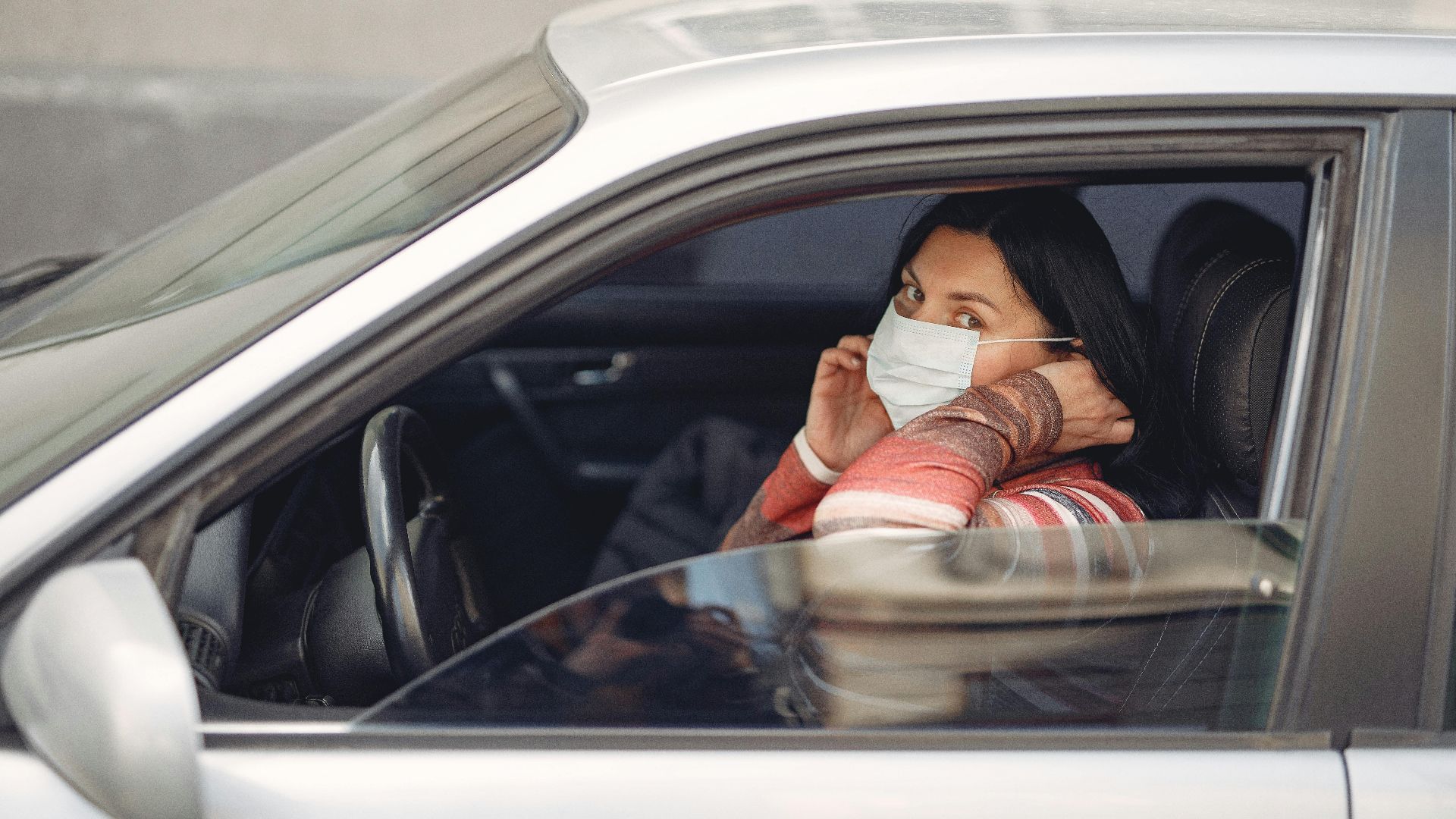 Female driver wearing warm outfit adjusting protective facial mask while sitting in auto with opened window during coronavirus pandemic in city and looking at camera