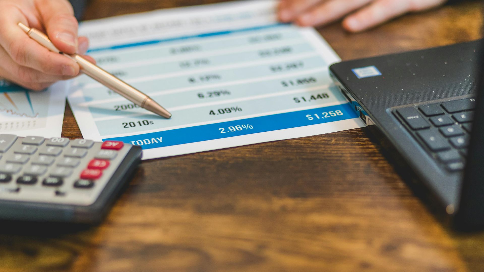 Close-up of a hand with pen analyzing financial rates on paper with a calculator and laptop nearby.