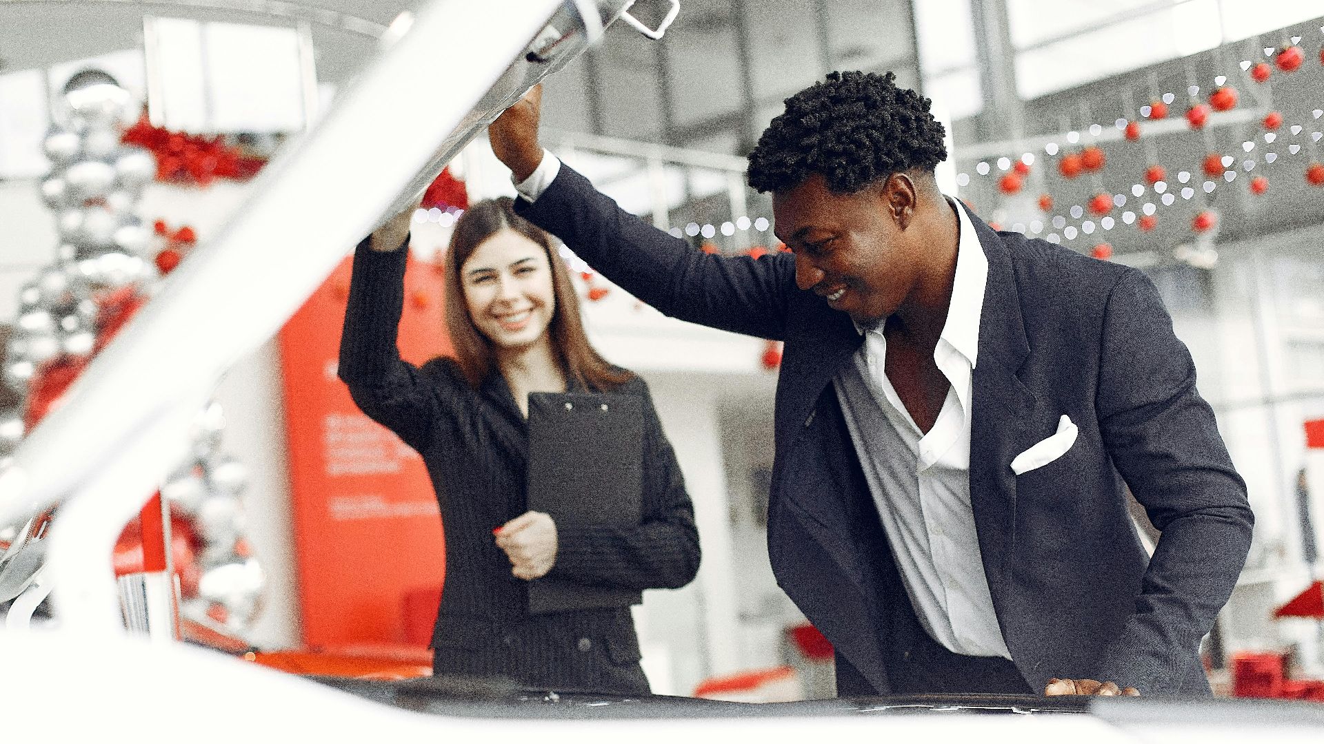 Positive smiling diverse stylish man and female dealer checking under car motor hood and smiling while choosing new car in car showroom