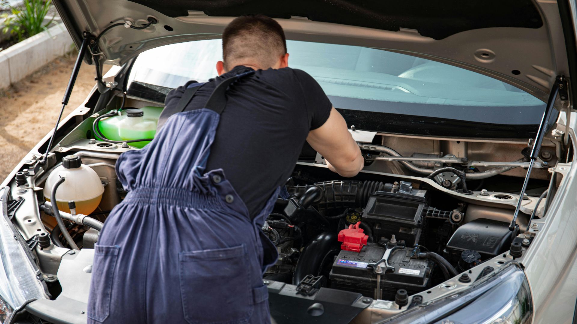 Mechanic in blue coveralls performing car engine maintenance outdoors.