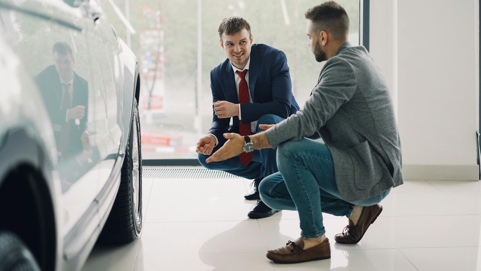Two men discussing car features in a showroom, kneeling near a vehicle.