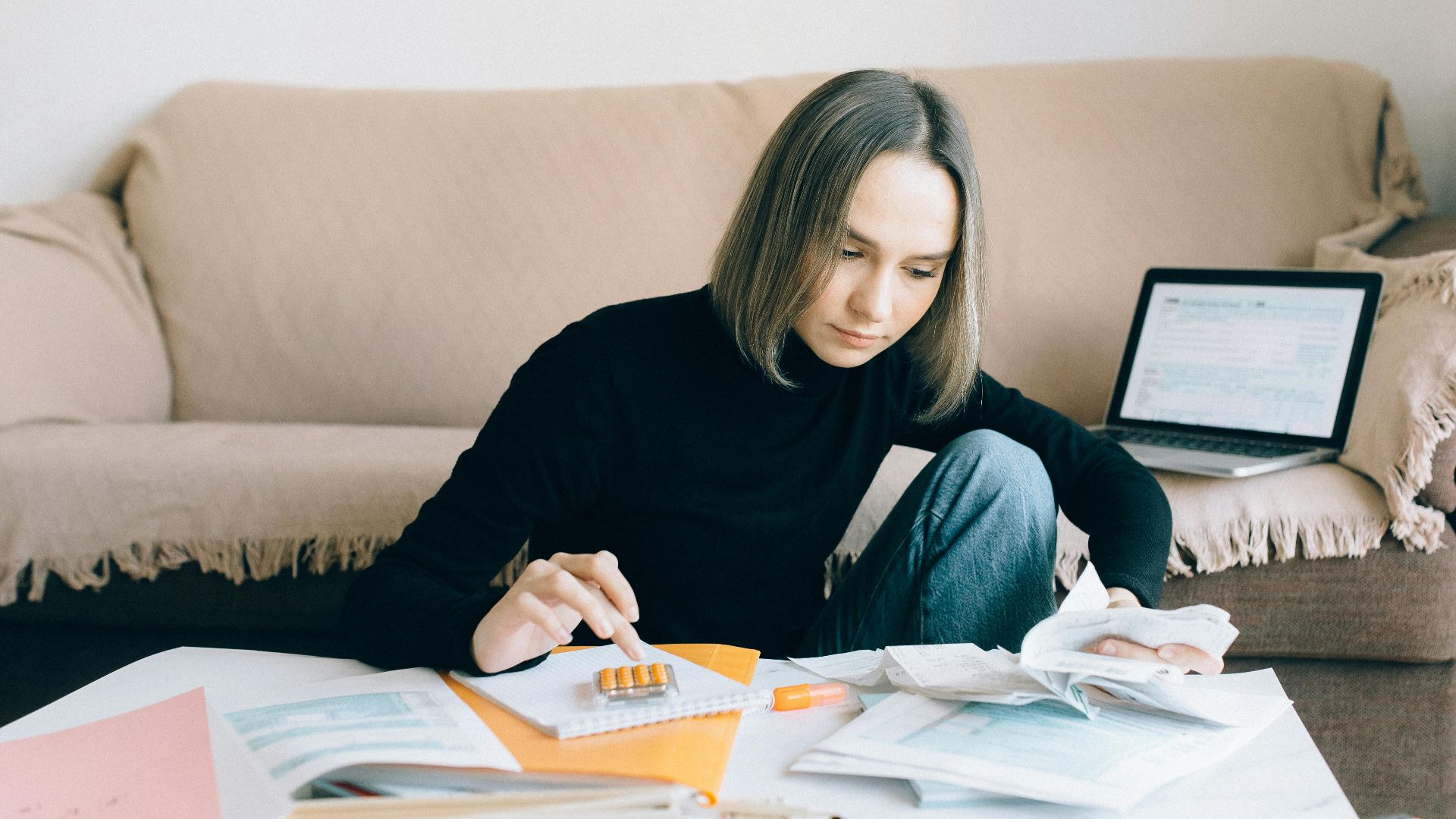 Young woman handling financial tasks with papers and laptop in cozy living room.