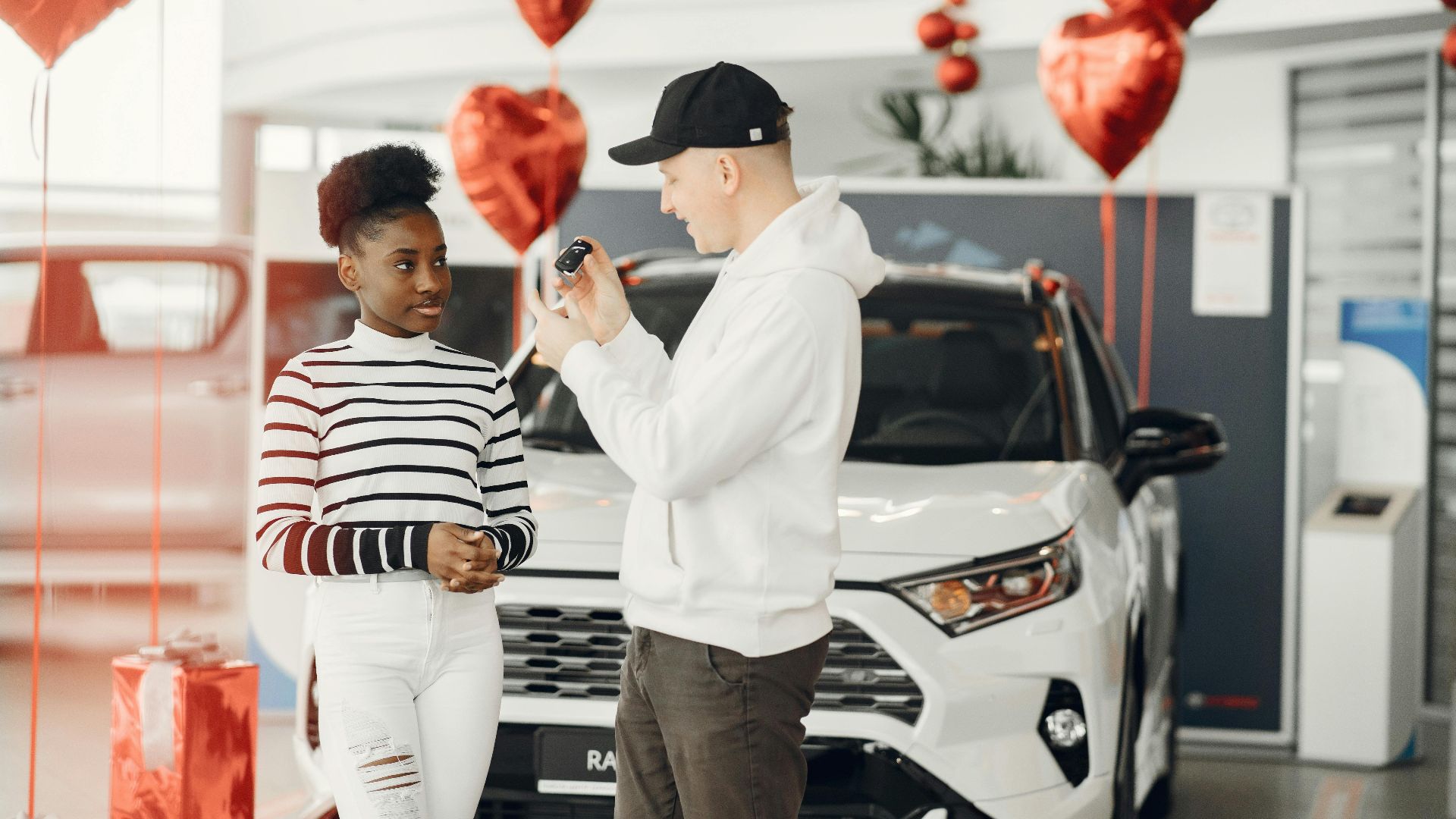 A couple exchanging car keys inside a showroom decorated with heart-shaped balloons, celebrating a new car purchase.