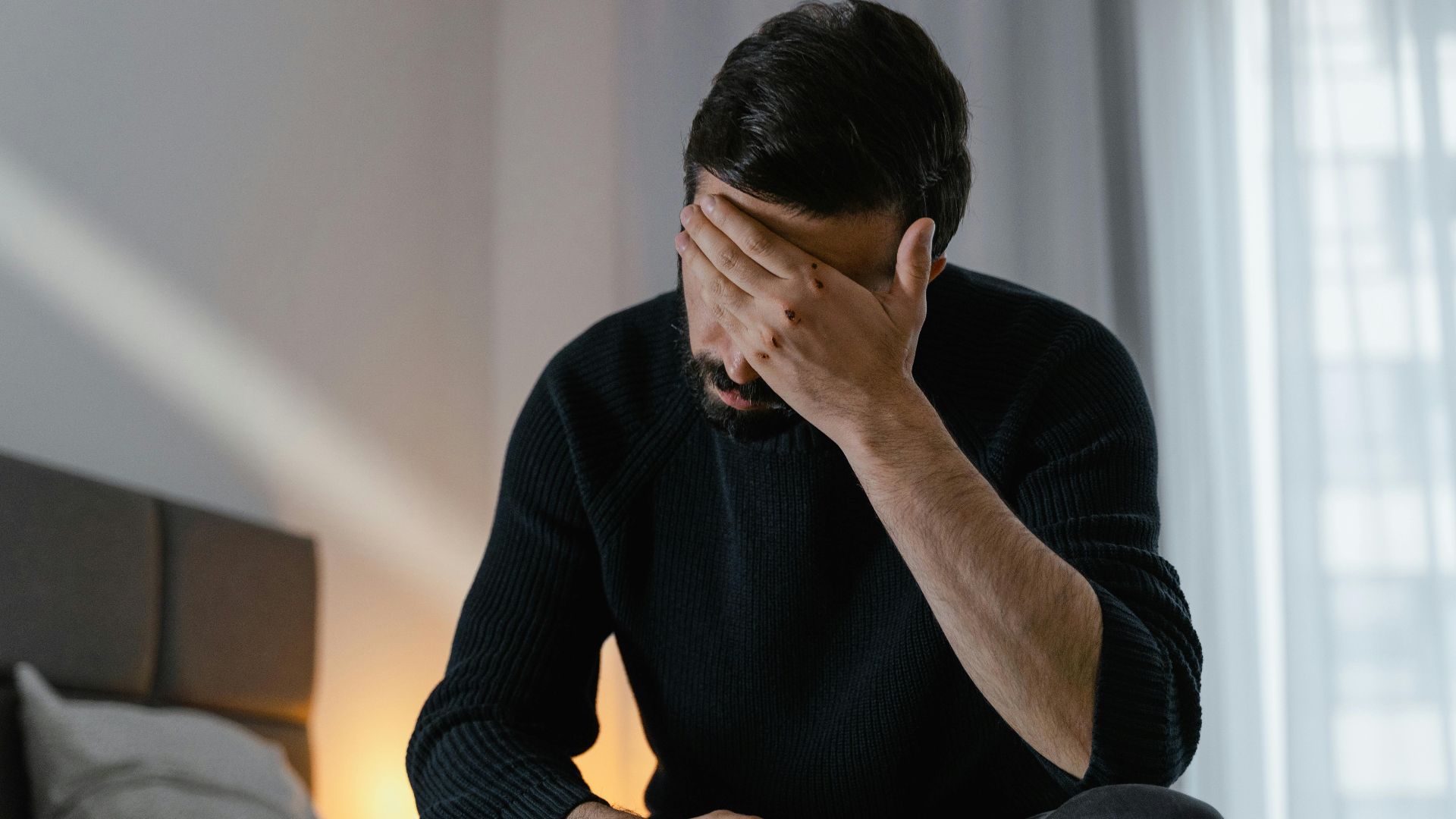 A man in distress sits on a bed indoors, covering his face with his hand.