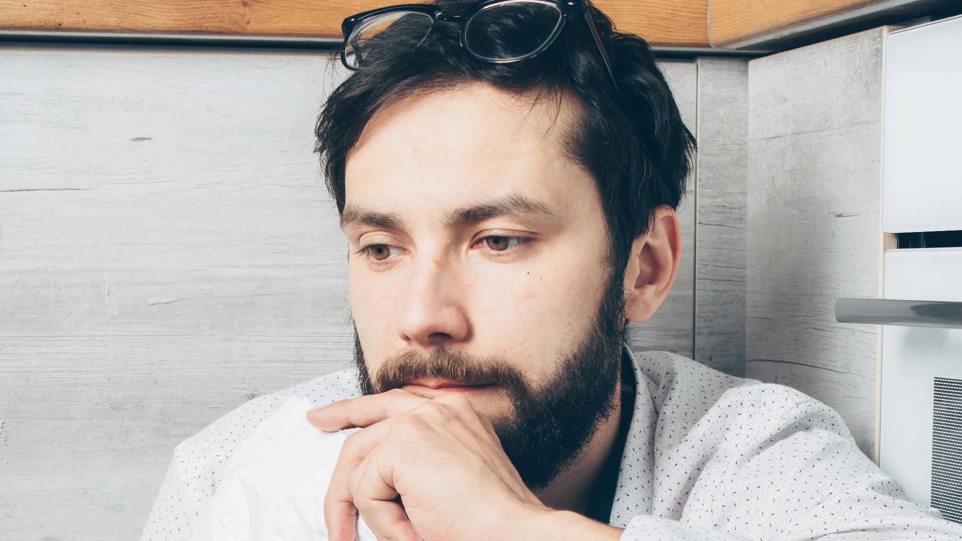 A young bearded man in deep thought holding a tissue, sitting indoors.