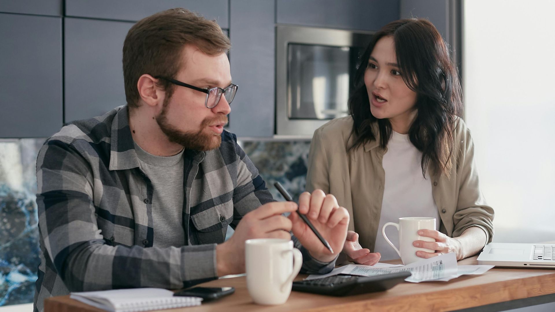 A couple managing finances with gadgets, documents, and coffee mugs at home.