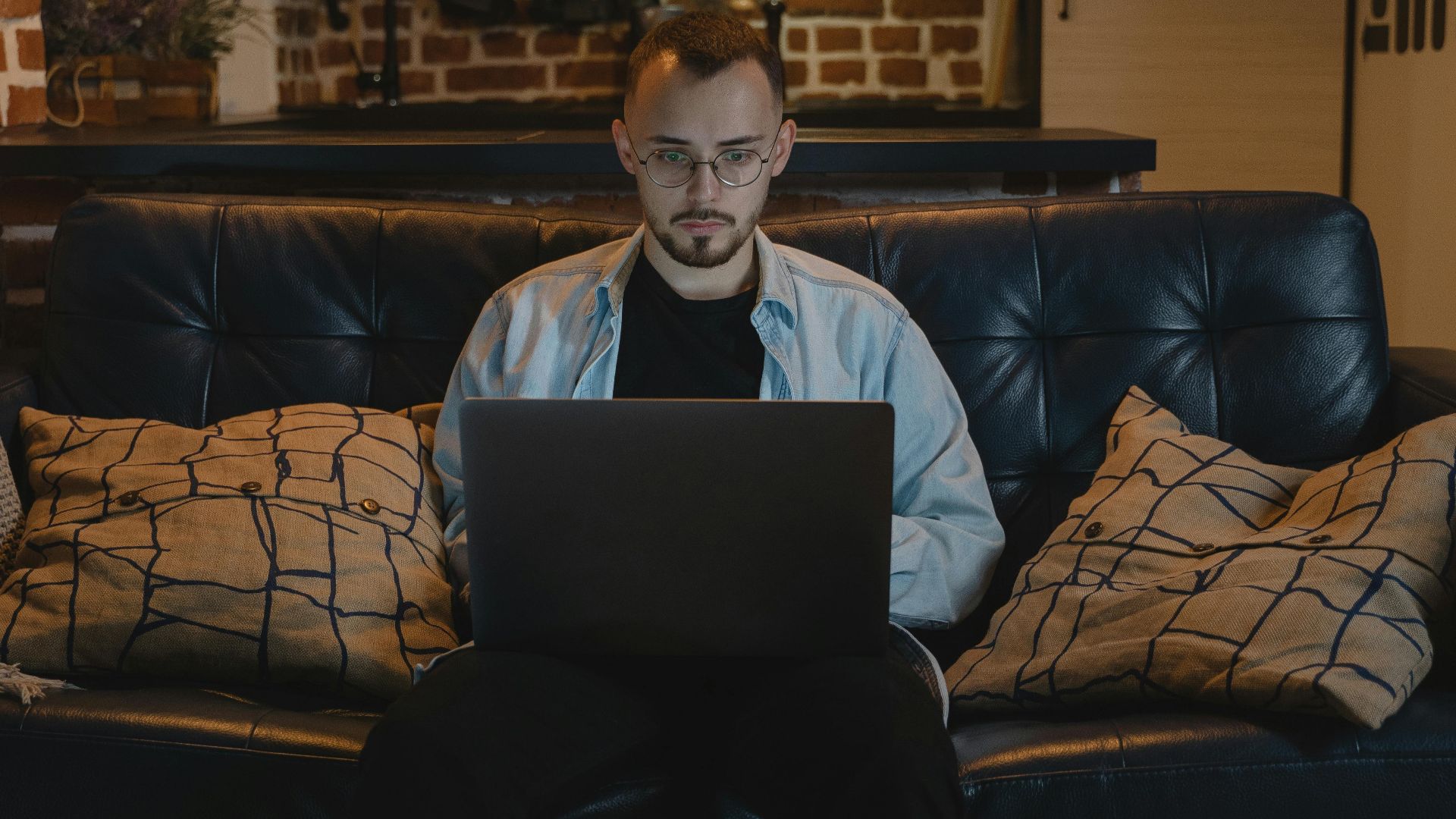 Man working remotely on a laptop in a cozy, stylish apartment with exposed brick walls and modern decor.