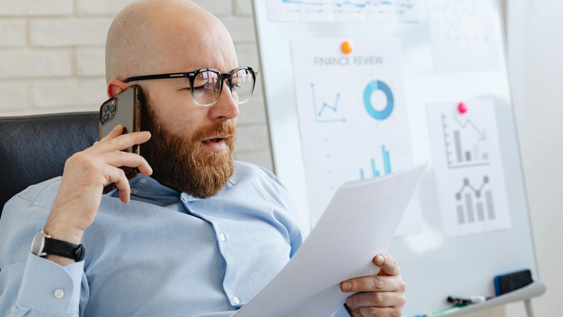 Bald man with beard holding smartphone and papers, reviewing financial charts in office.