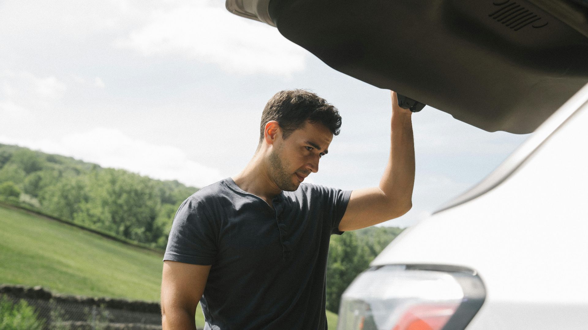 A man examining the open trunk of a car under clear skies with greenery in the background.
