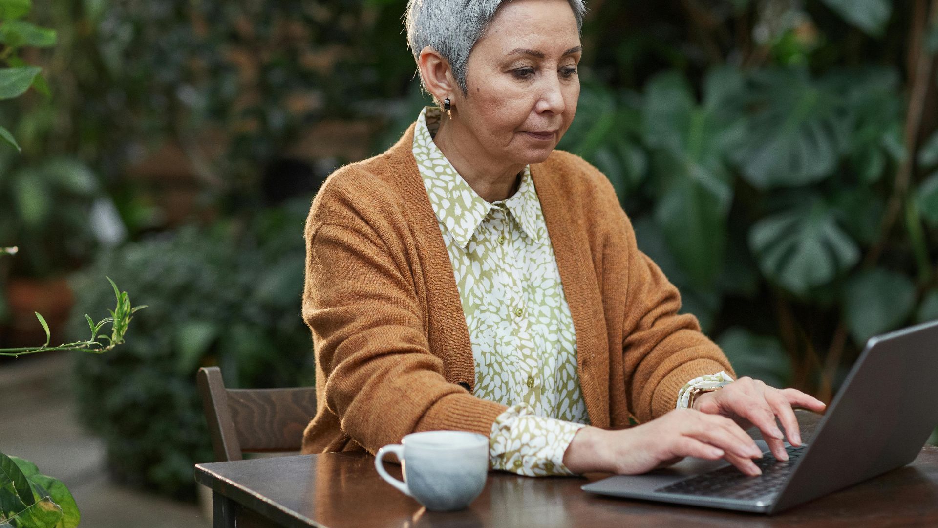 An elderly woman using a laptop outdoors surrounded by lush green plants.