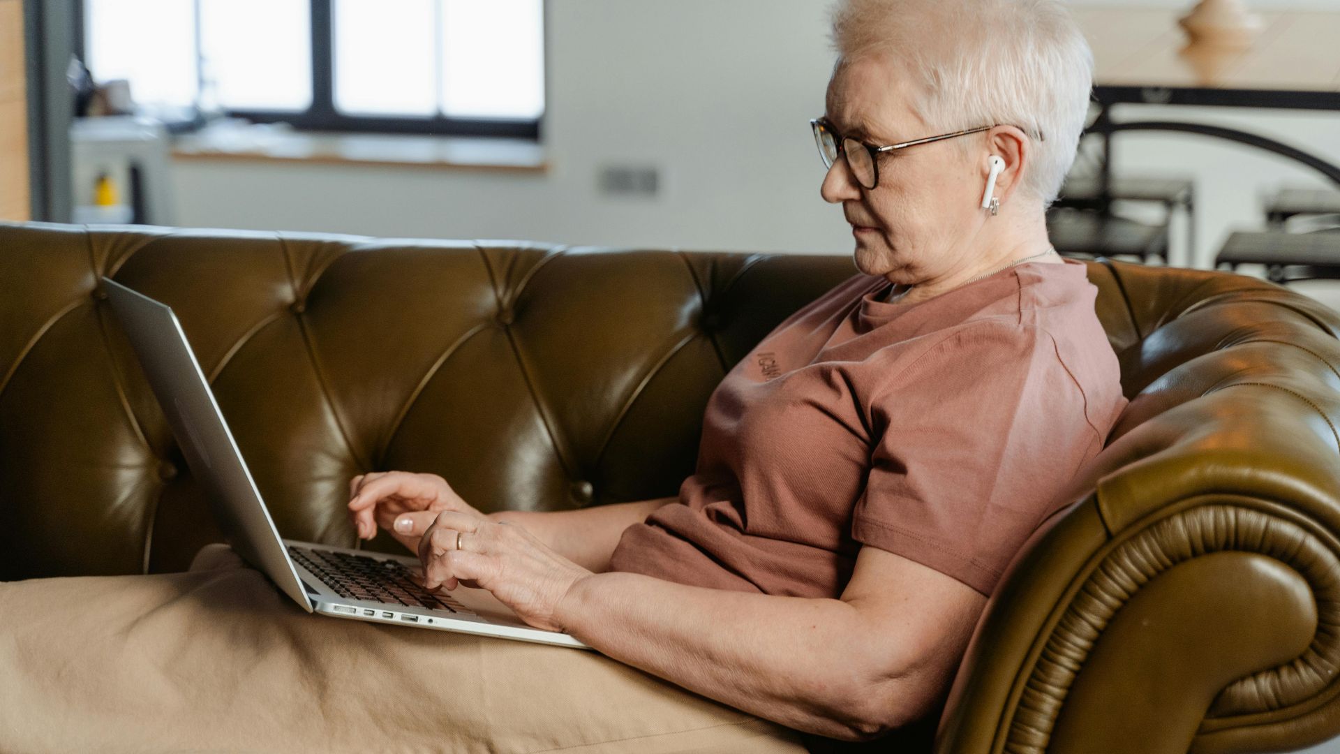 Elderly woman relaxing on a couch, using a laptop indoors during the day.