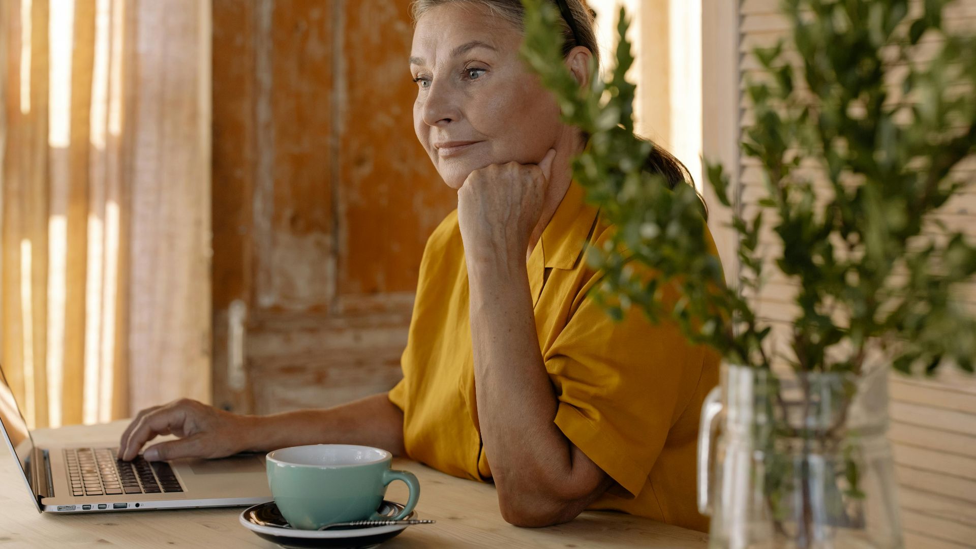 Elderly woman sitting at a table using a laptop, relaxing in a cozy home setting.