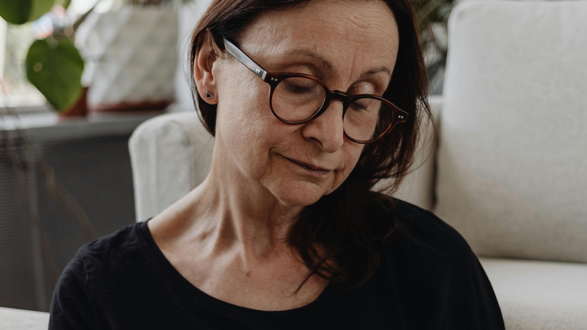 Elderly woman in glasses sitting reflectively indoors, surrounded by plants.