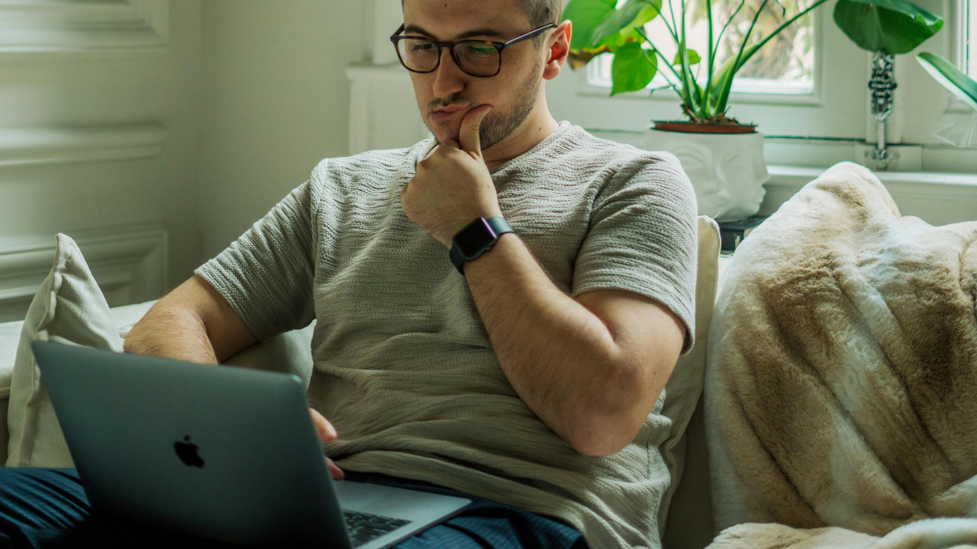A man wearing eyeglasses using a laptop in a cozy home setting with natural light.