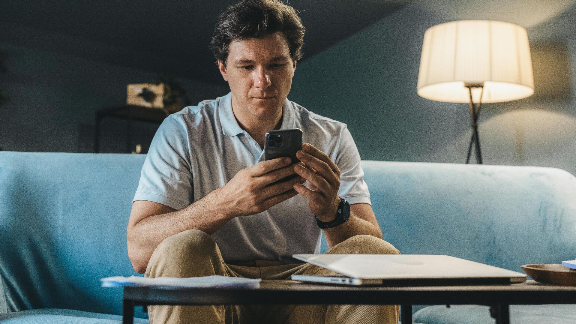 A man sitting on a sofa indoors using a smartphone under warm lighting.