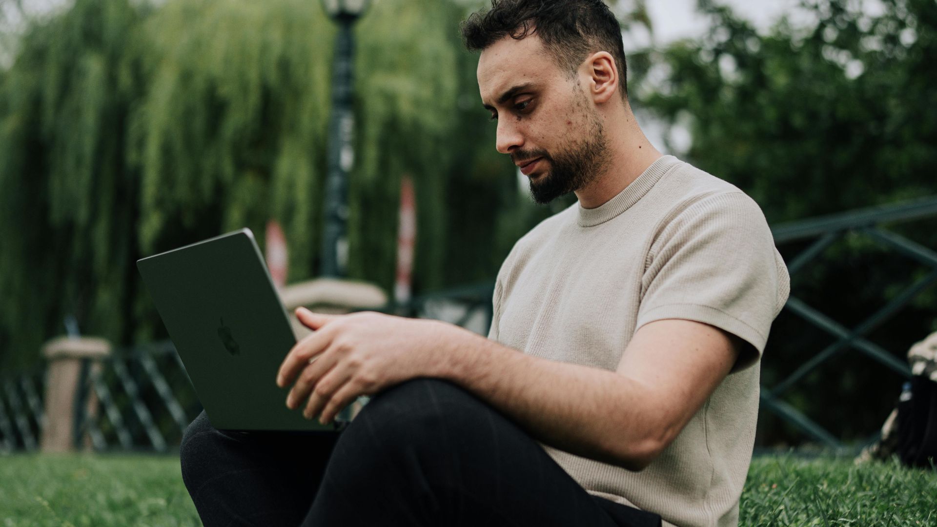 Casual man sitting on grass in park, browsing on laptop during a summer day.