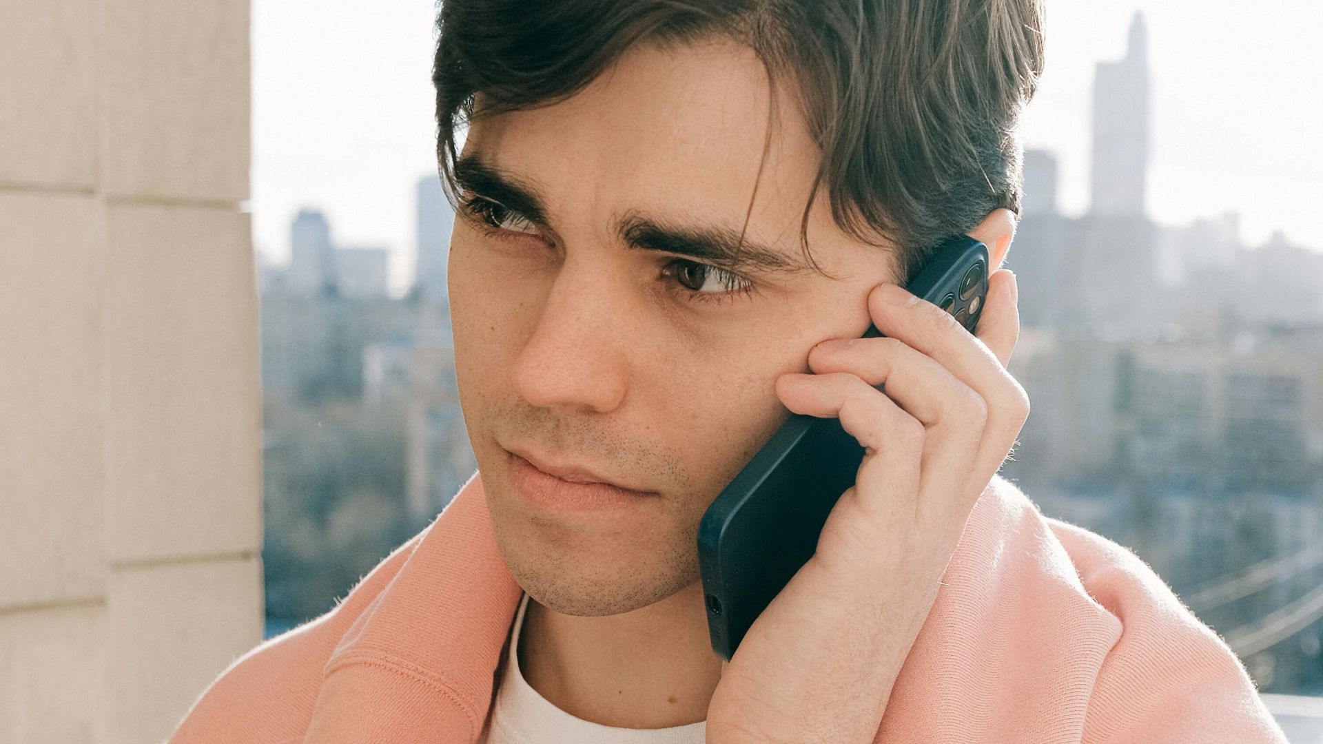 Young man talks on cellphone on an urban balcony with city skyline in the background.