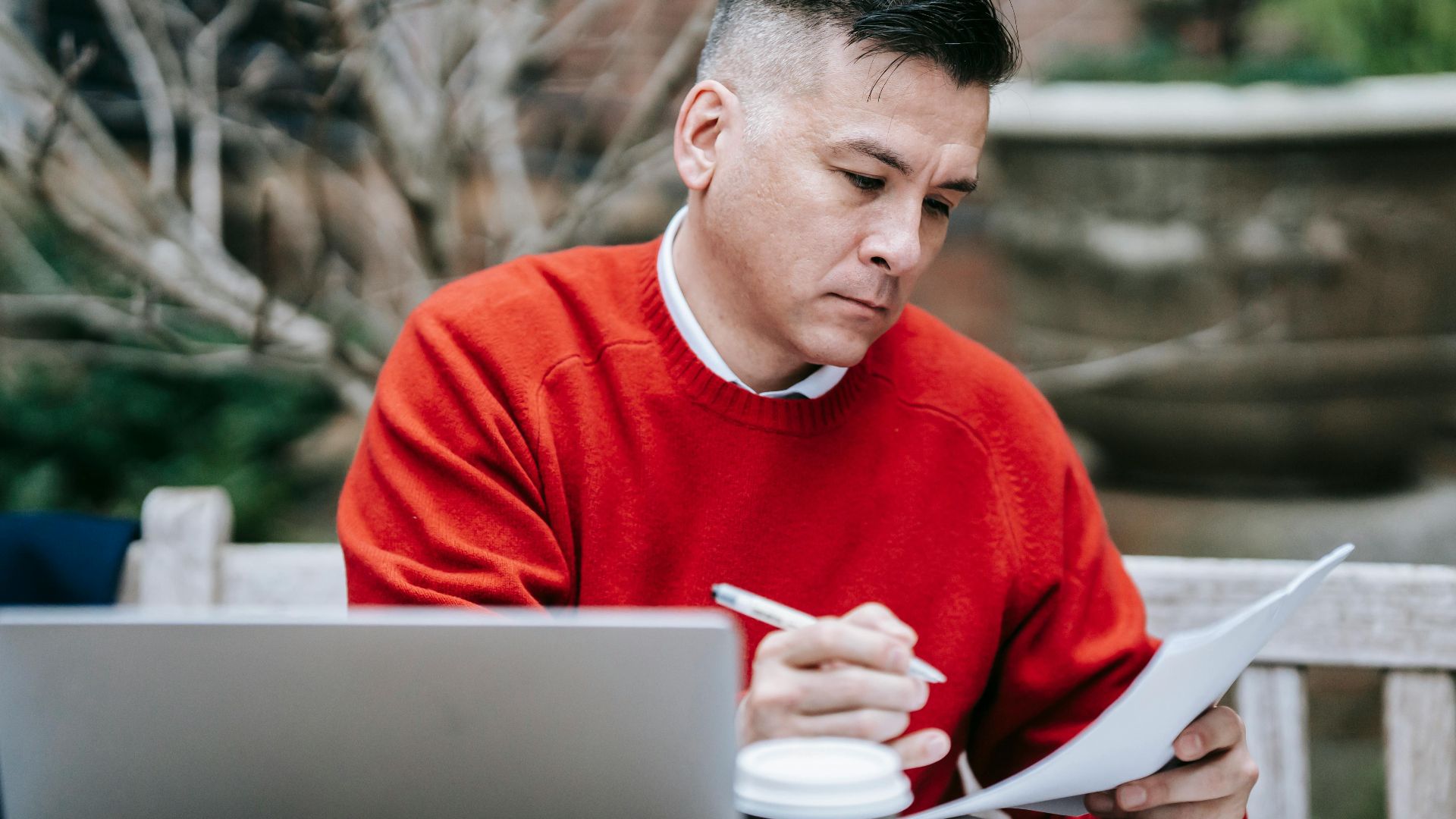 A man in a red sweater concentrating on paperwork while working remotely with a laptop outdoors.