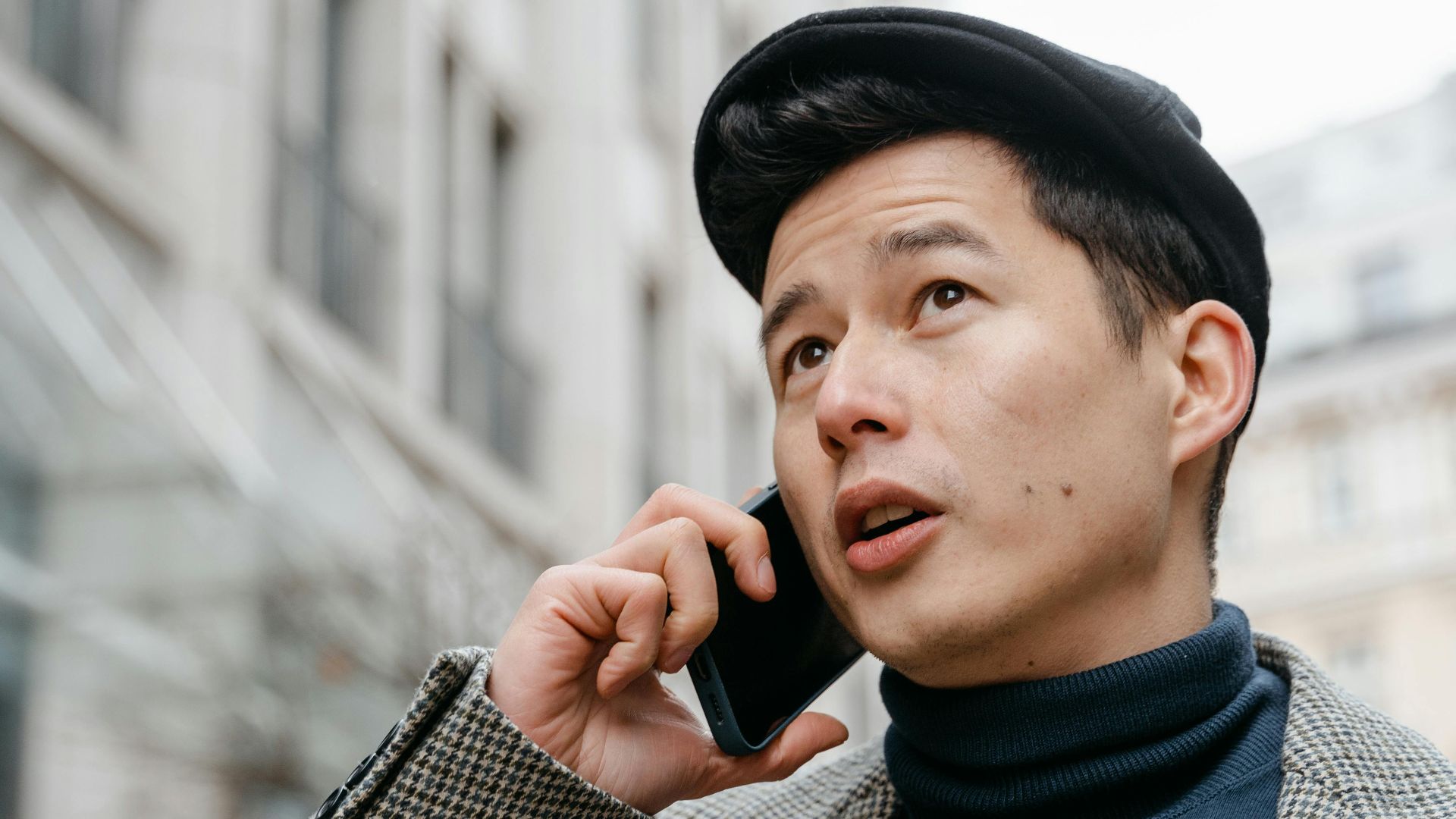 Stylish man in a city talking on a smartphone outdoors, wearing a coat and hat.