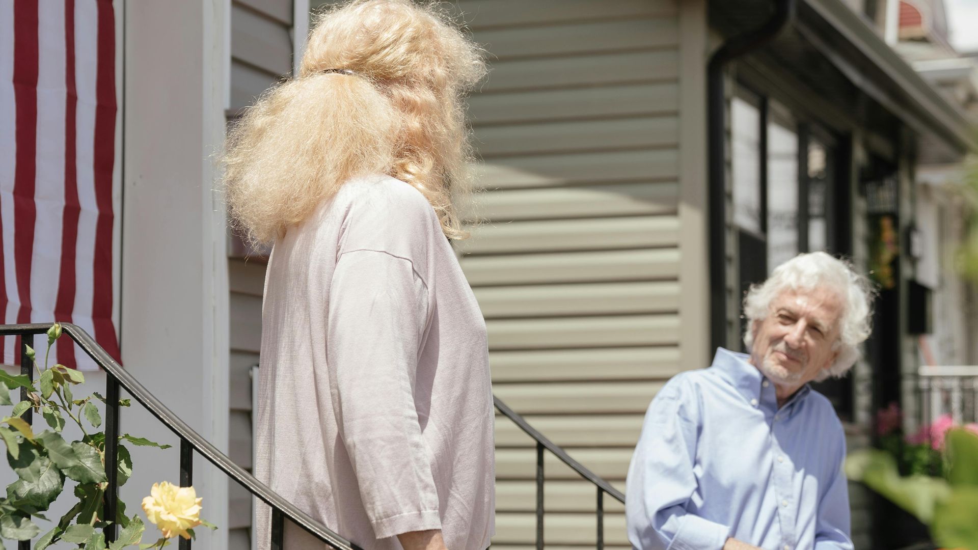 An elderly couple engaging in conversation on the porch of their suburban home with an American flag displayed.