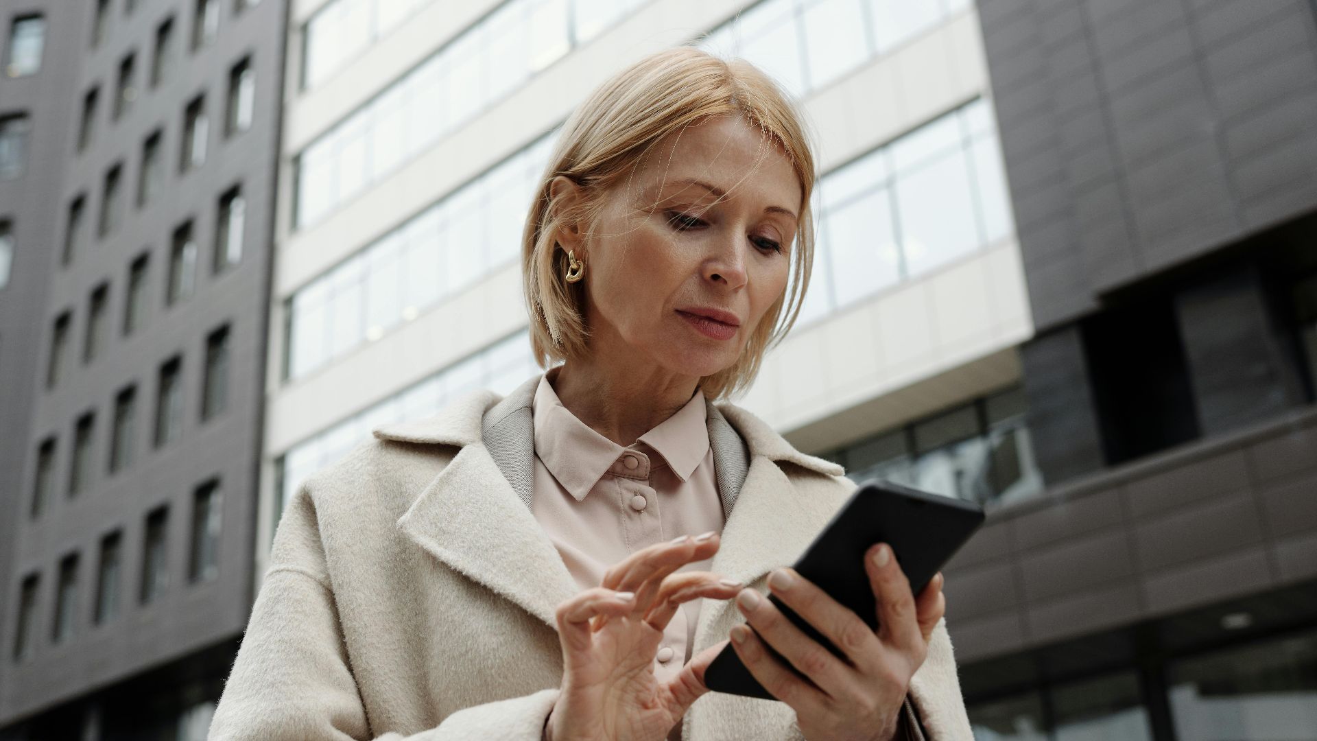 Woman in coat using smartphone in front of modern building. Professional and focused expression.