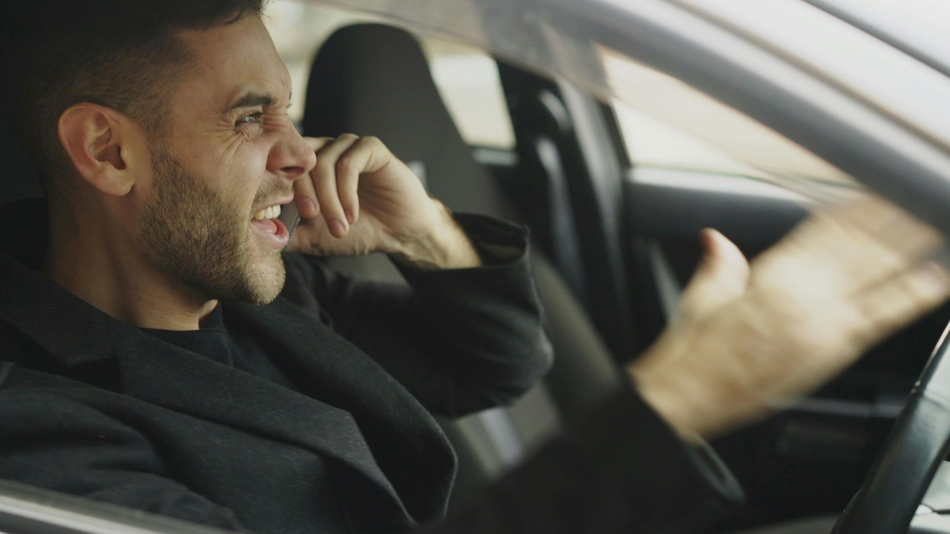 An adult man expressing frustration while on the phone inside a car.