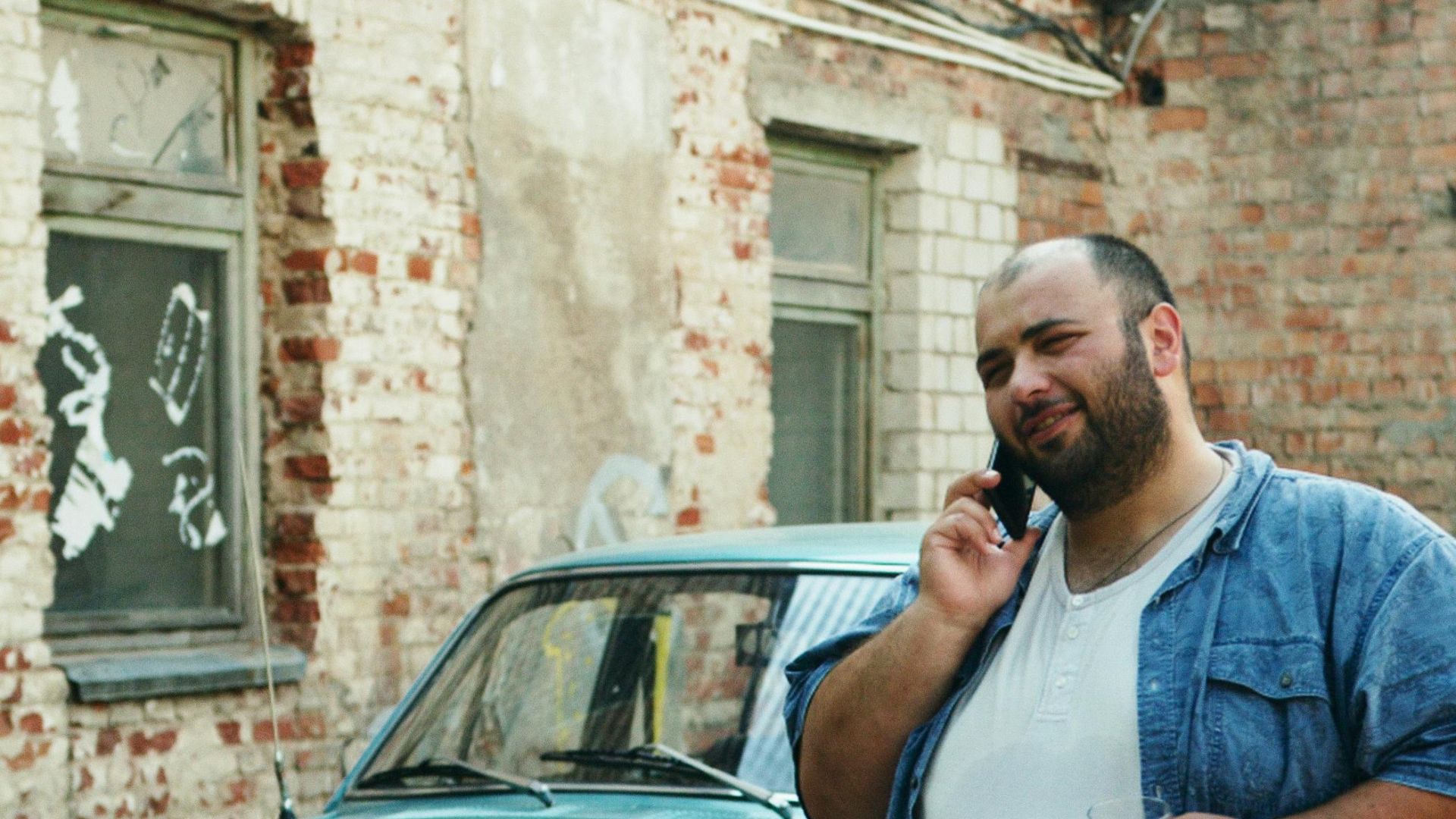 Casually dressed man talking on the phone next to a vintage car in an urban setting.