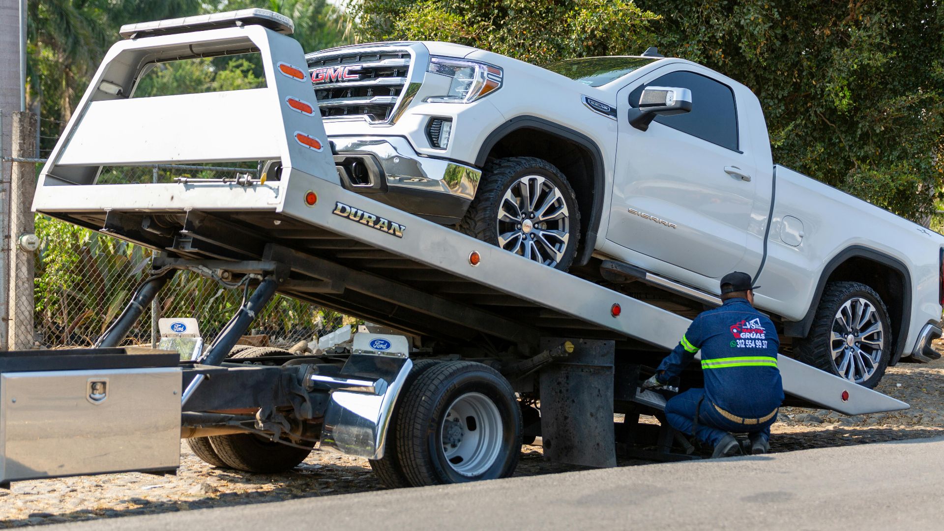 Tow truck operator loading white GMC pickup truck on street in daytime.