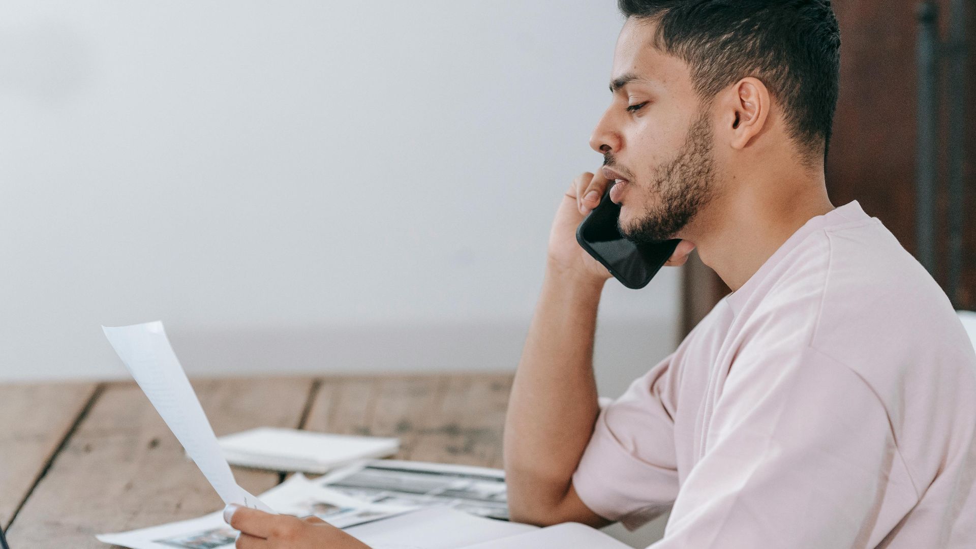 Side view of young Hispanic male remote employee discussing documents during phone call while working online with laptop and papers in home office