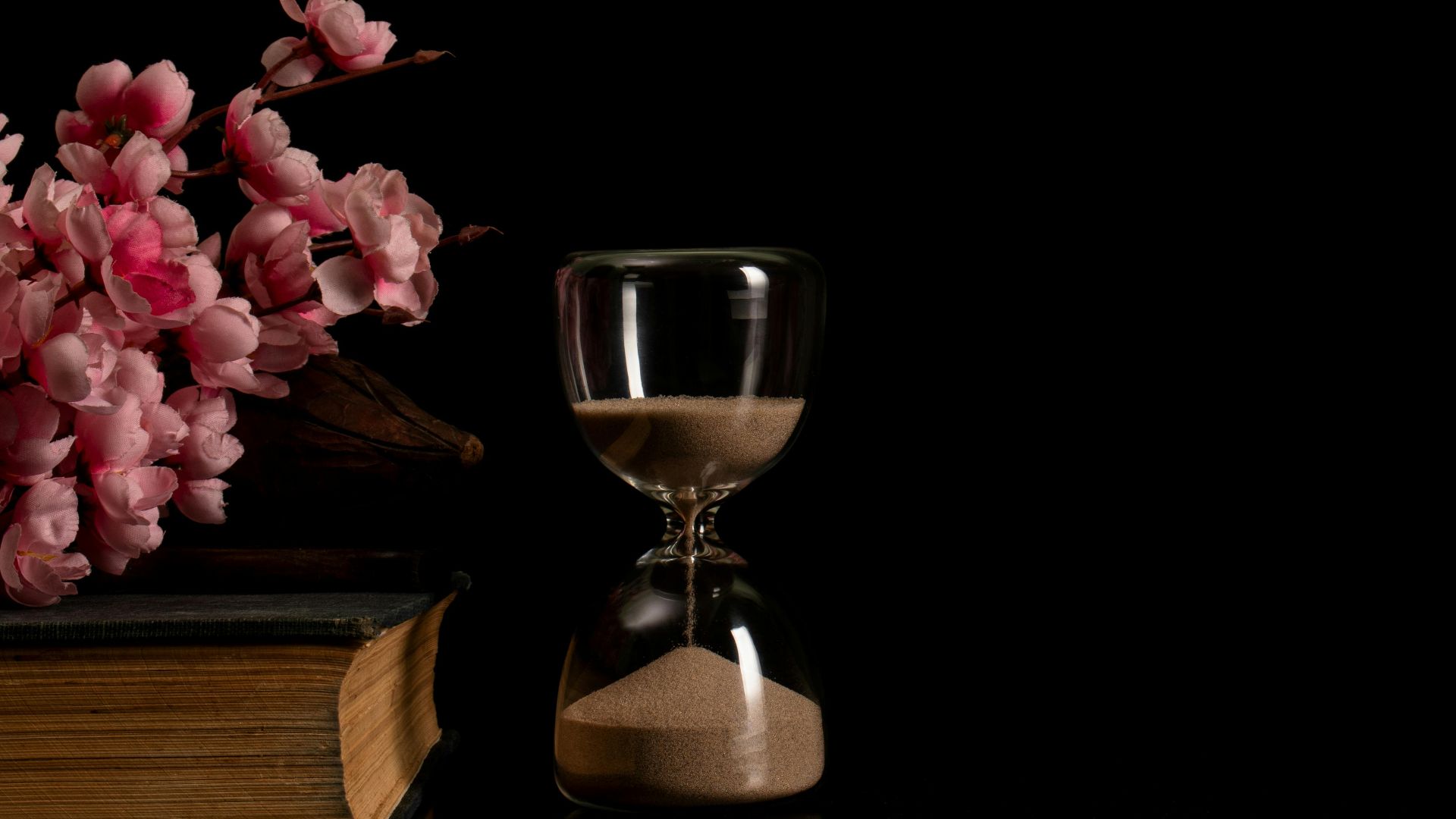 Charming still life with pink flowers, vintage book, and hourglass on a black background.