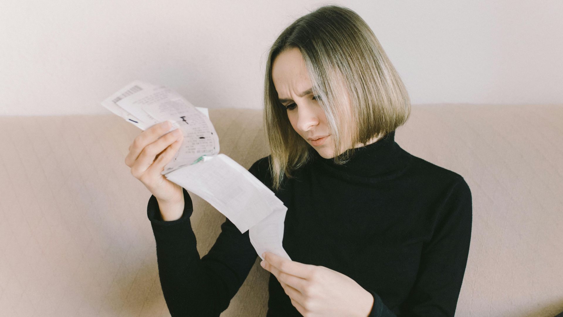 Concerned woman in black sweater examining bills on beige sofa indoors.