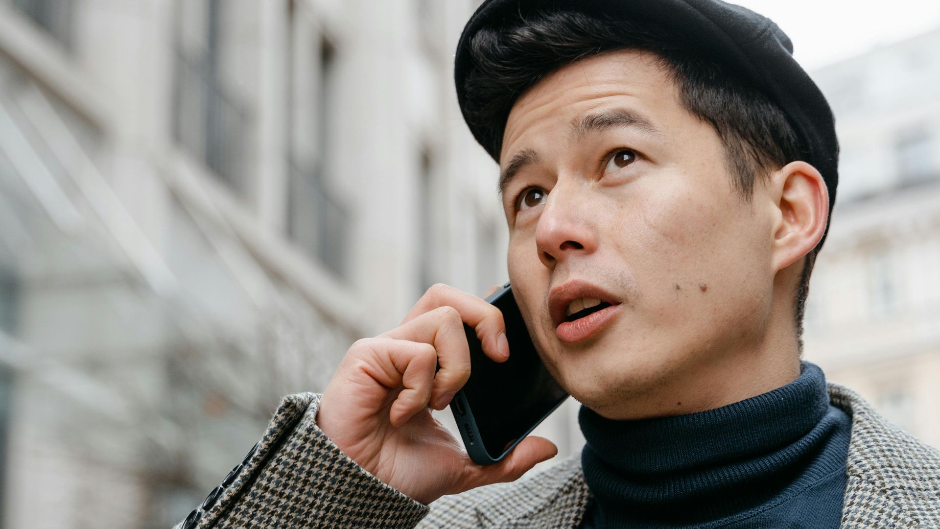 Stylish man in a city talking on a smartphone outdoors, wearing a coat and hat.