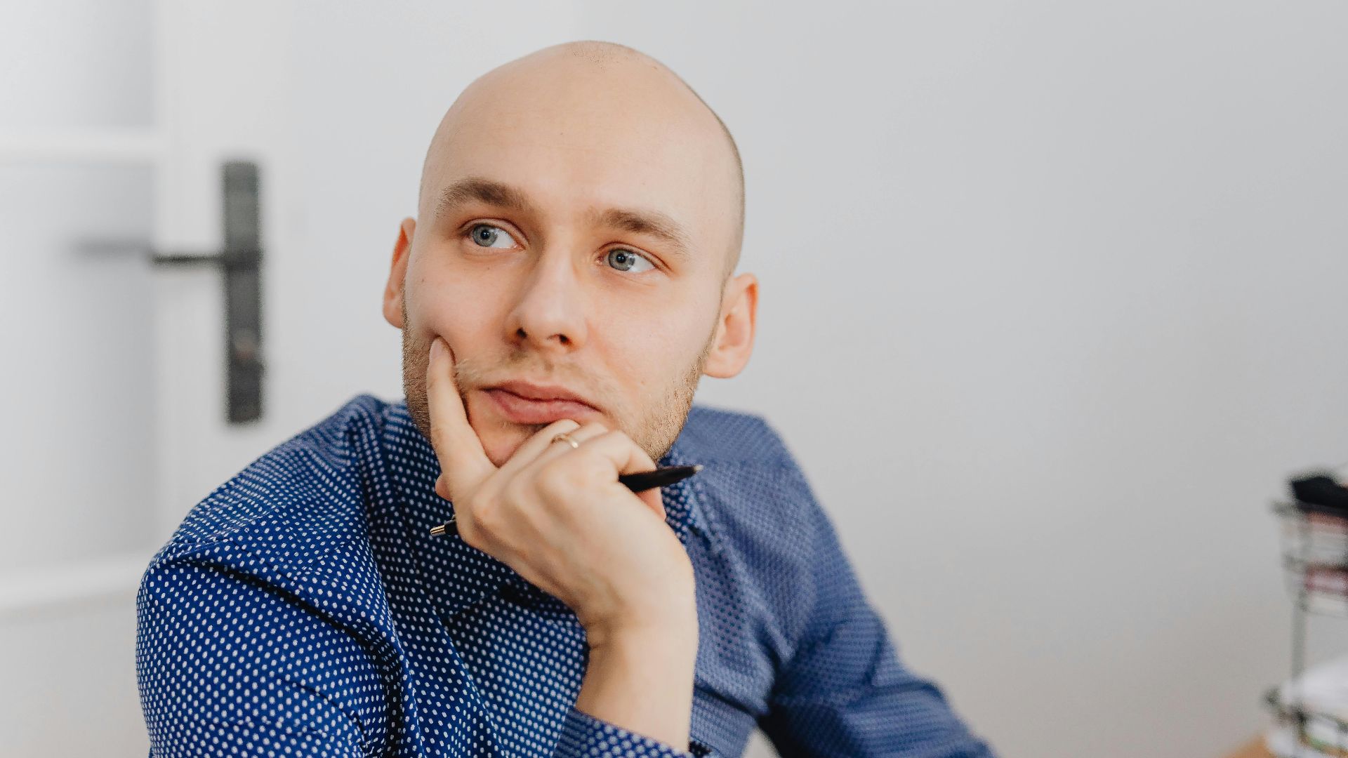 Bald man in blue shirt thoughtfully holding a pen indoors, gazing into distance.