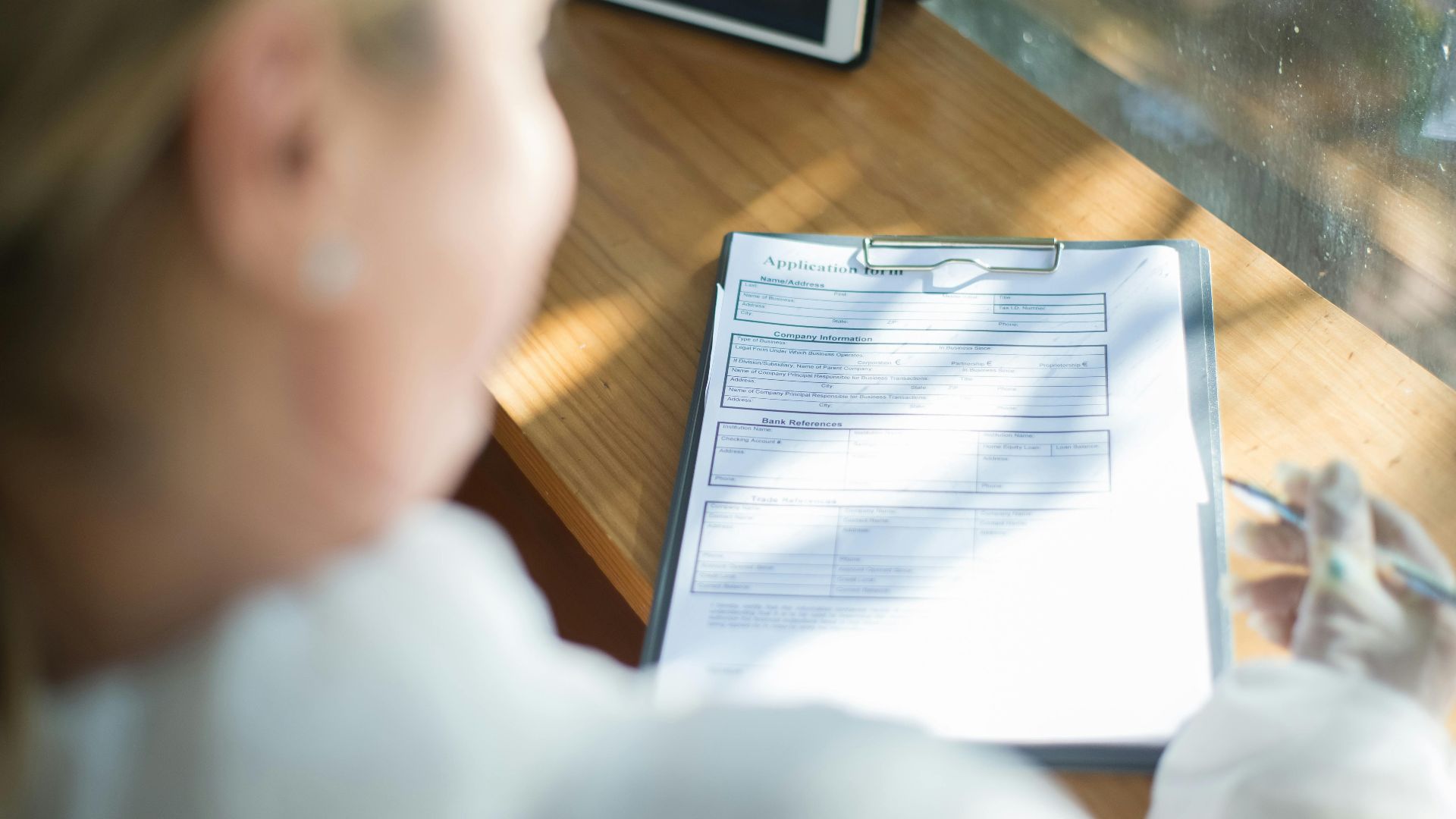 A woman fills out an application form with a pen at a sunlit wooden desk.