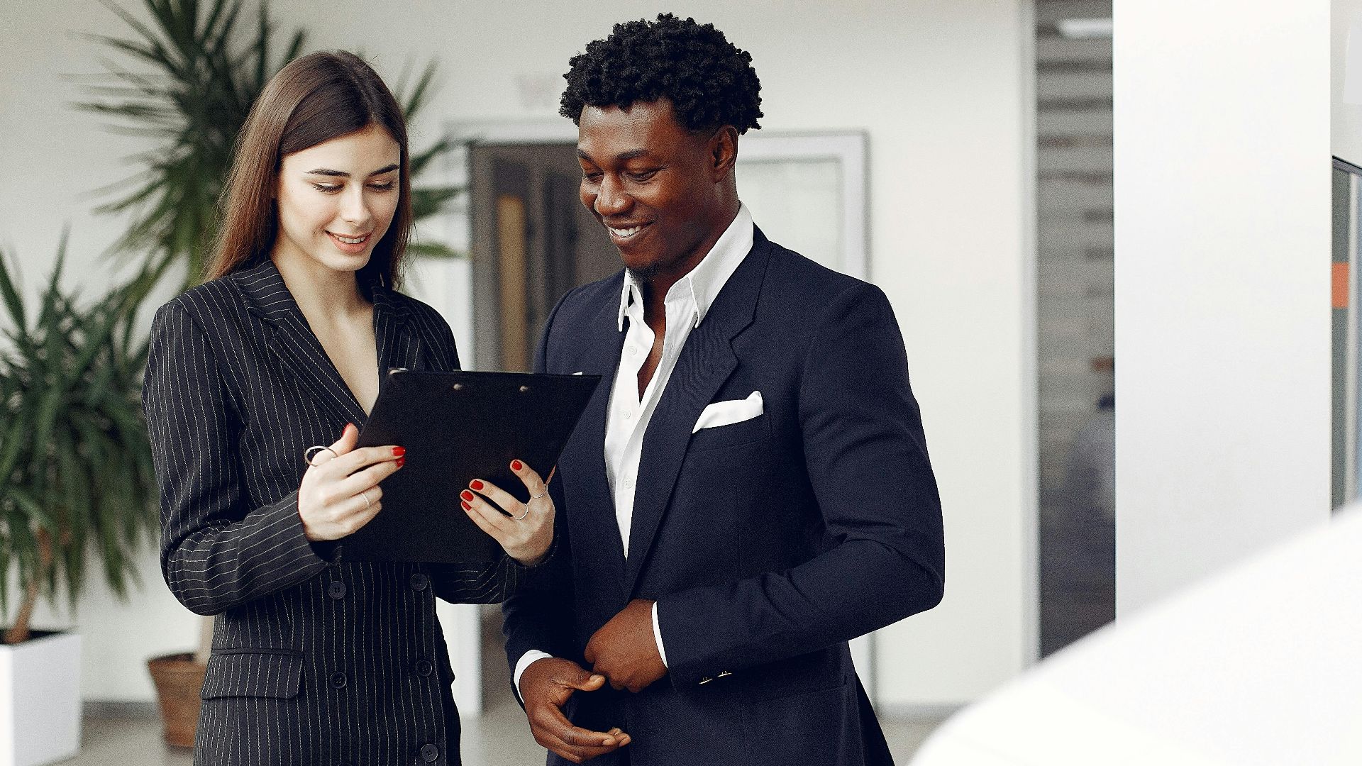 Cheerful multiethnic stylish female agent and smiling customer in formal suit discussing contract details while standing in modern car showroom in daylight