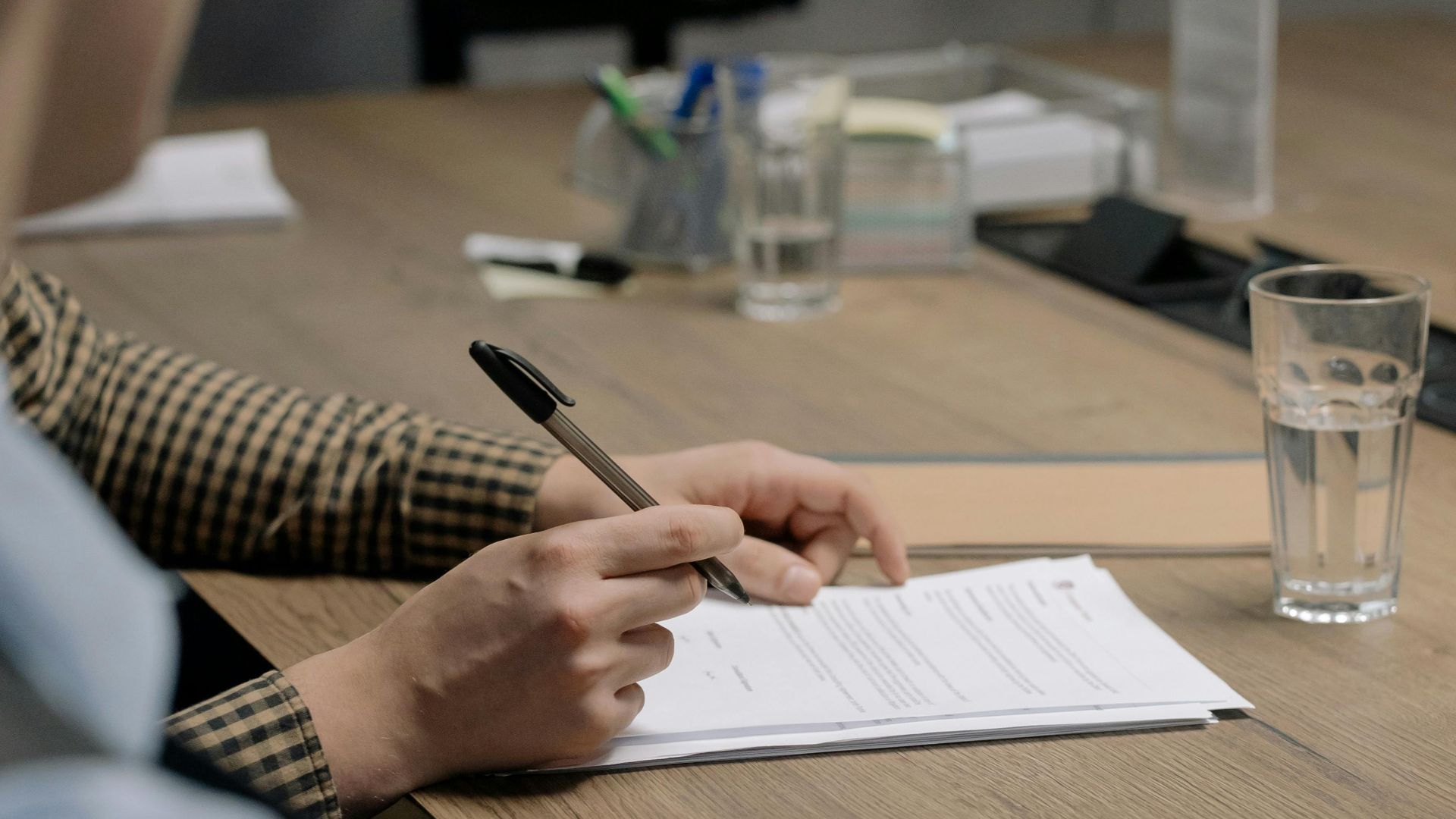 Person in business attire signing a document at a wooden table in an office setting.