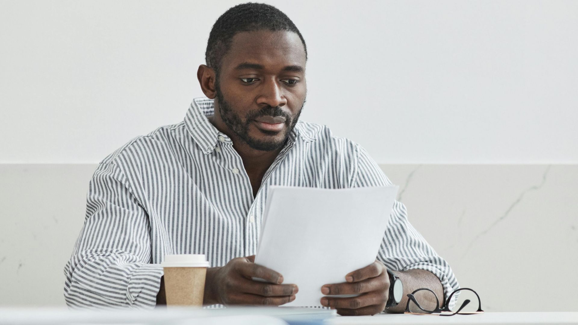 African American man sitting indoors, reading papers with a coffee cup nearby.